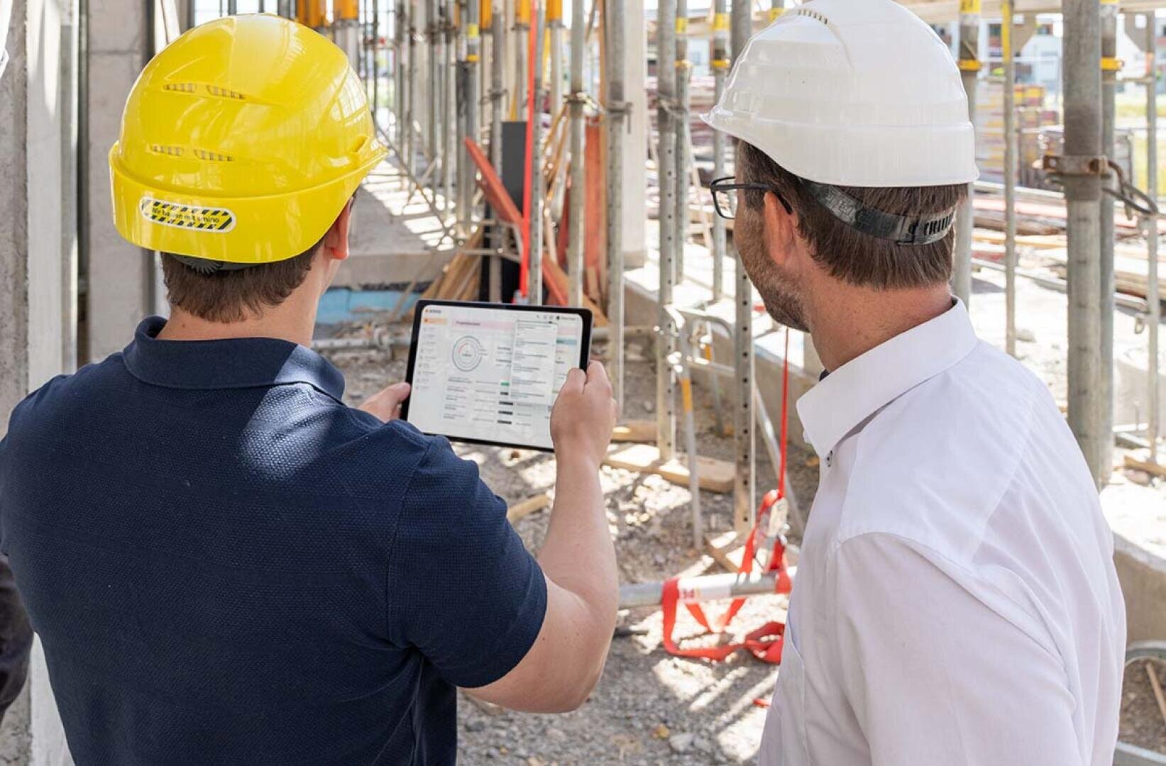 Zwei Bauarbeiter mit Schutzhelmen schauen auf ein Tablet, das Daten auf einer Baustelle mit Gerüsten und Baumaterialien im Hintergrund anzeigt.