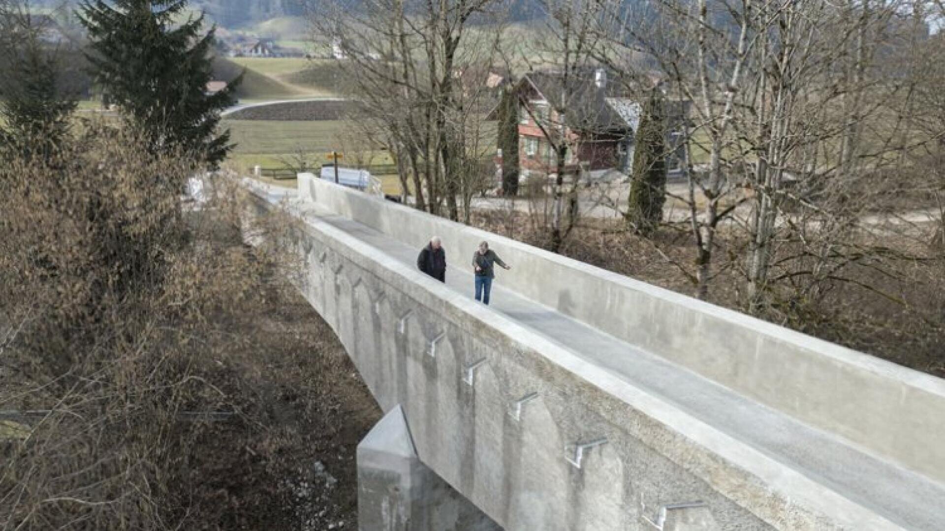 Zwei Menschen gehen über eine breite, erhöhte Fußgängerbrücke aus Beton, umgeben von Bäumen und Landschaft, im Hintergrund sind Häuser und Felder zu sehen.