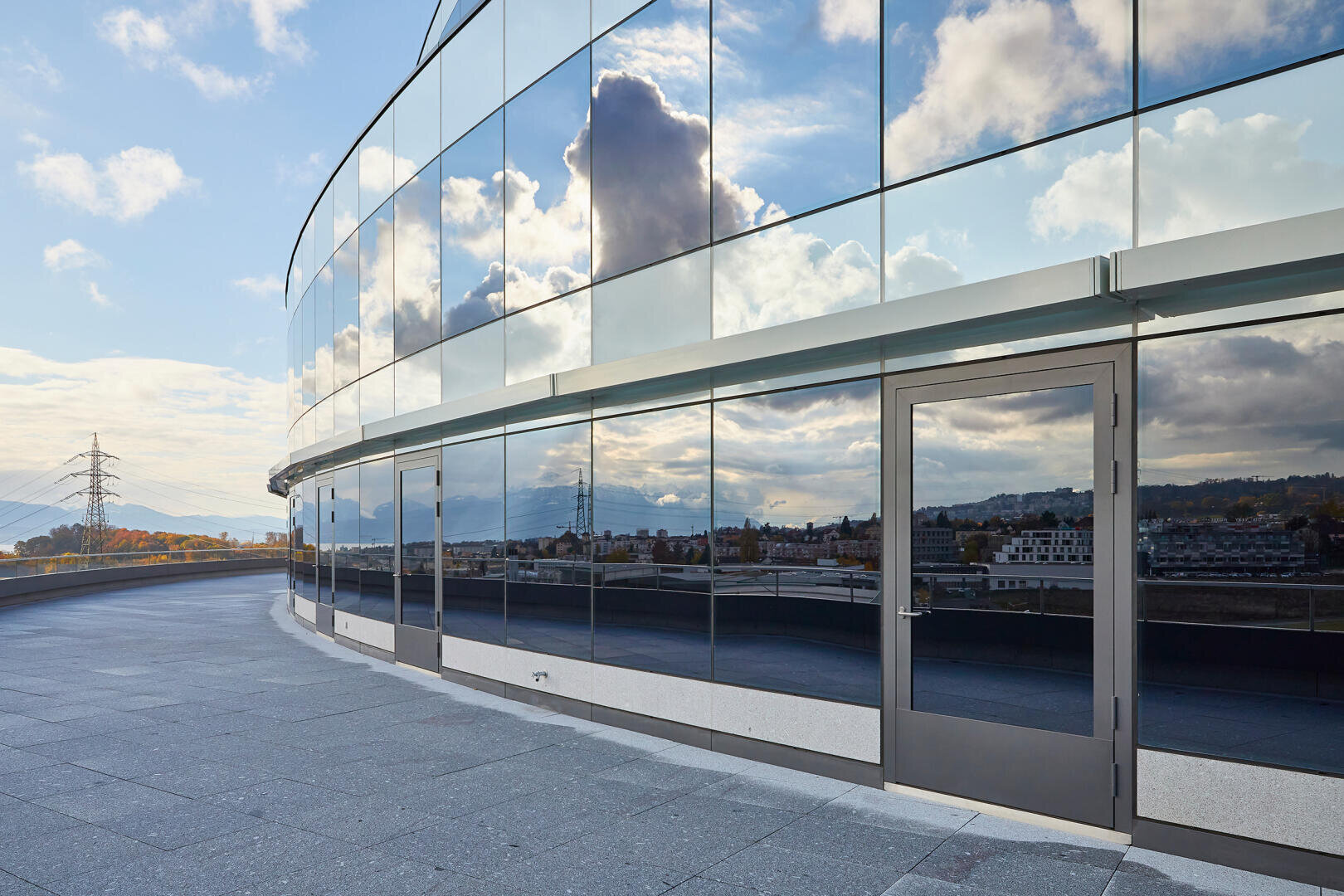 Ein modernes Glasgebäude mit spiegelnden Fenstern schlängelt sich auf einem gepflasterten Gehweg unter einem teilweise bewölkten Himmel. Das Stadtbild, die Wolken und die Stromleitungen spiegeln sich im Glas.
