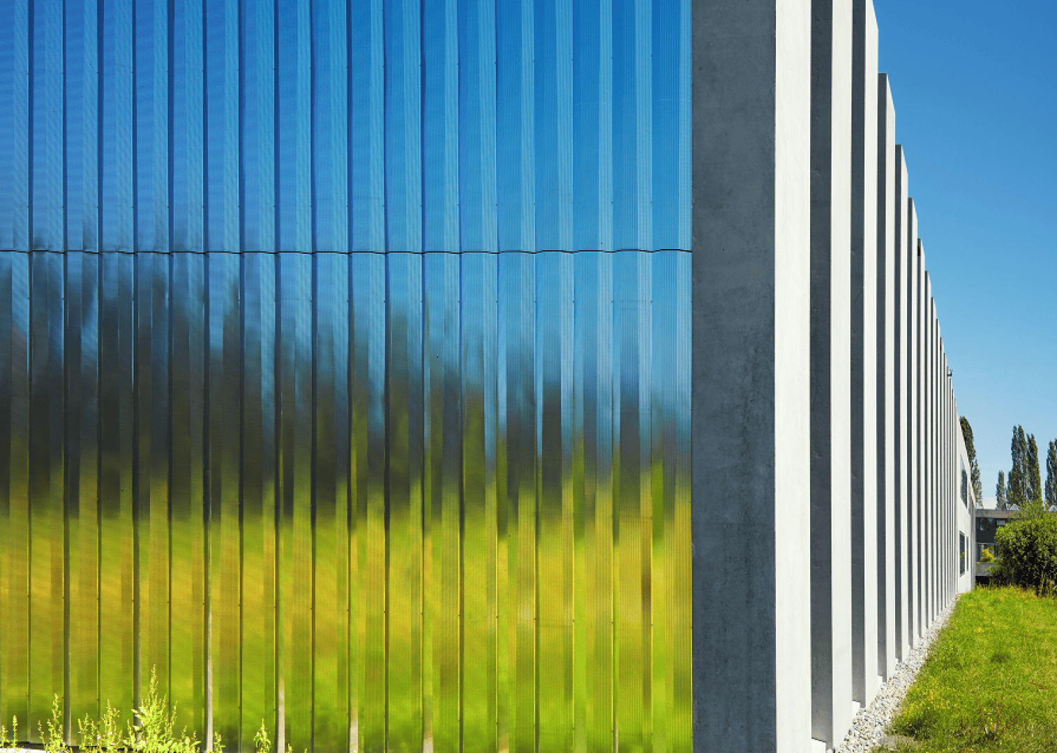 Ein modernes Gebäude mit hohen, vertikalen, reflektierenden Glaspaneelen neben Betonsäulen, in denen sich grünes Gras und blauer Himmel im hellen Sonnenlicht spiegeln.