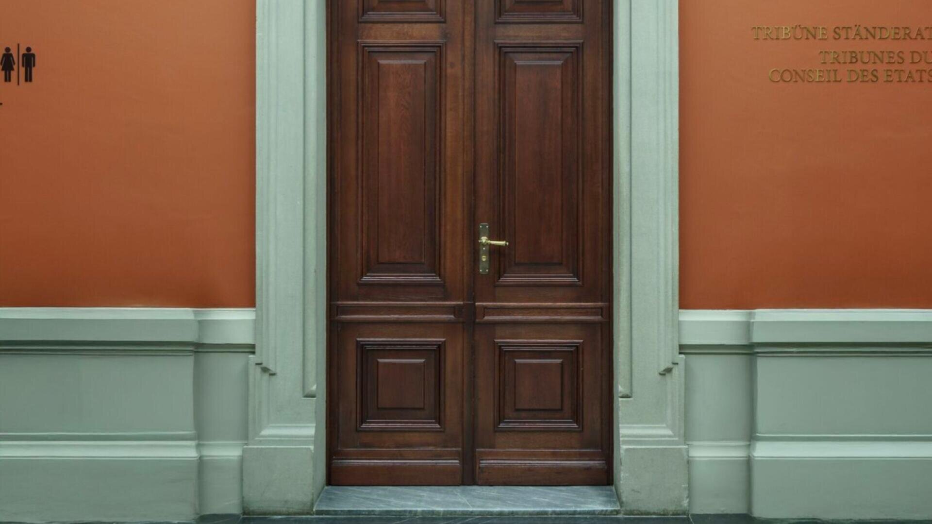 Une grande double porte en bois avec une poignée en laiton dans un cadre en pierre devant un mur orange avec des panneaux et des symboles de toilettes de chaque côté. Le mur a une partie inférieure blanche.