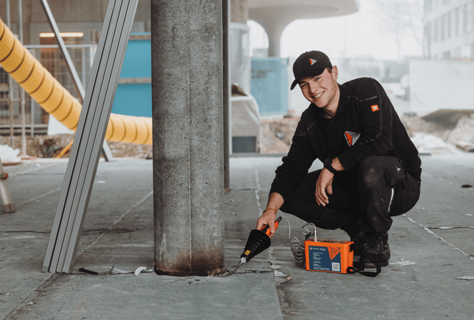 Ein Bauarbeiter in schwarzer Kleidung und einer Mütze kniet auf dem Boden und lächelt, während er ein Elektrowerkzeug an einer Metallsäule auf einer Baustelle mit Materialien und Geräten im Hintergrund benutzt.
