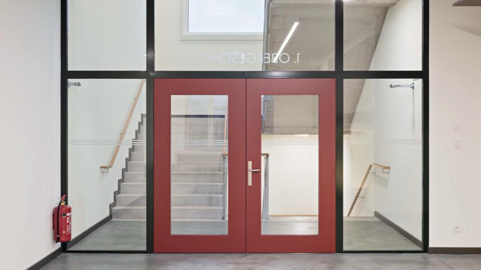 Ein modernes Treppenhaus mit einer roten Doppeltür und großen Glasscheiben, die zu einer Treppe führen. An der linken Wand ist ein Feuerlöscher angebracht, und über der Tür befindet sich ein Fenster, das Tageslicht hereinlässt.