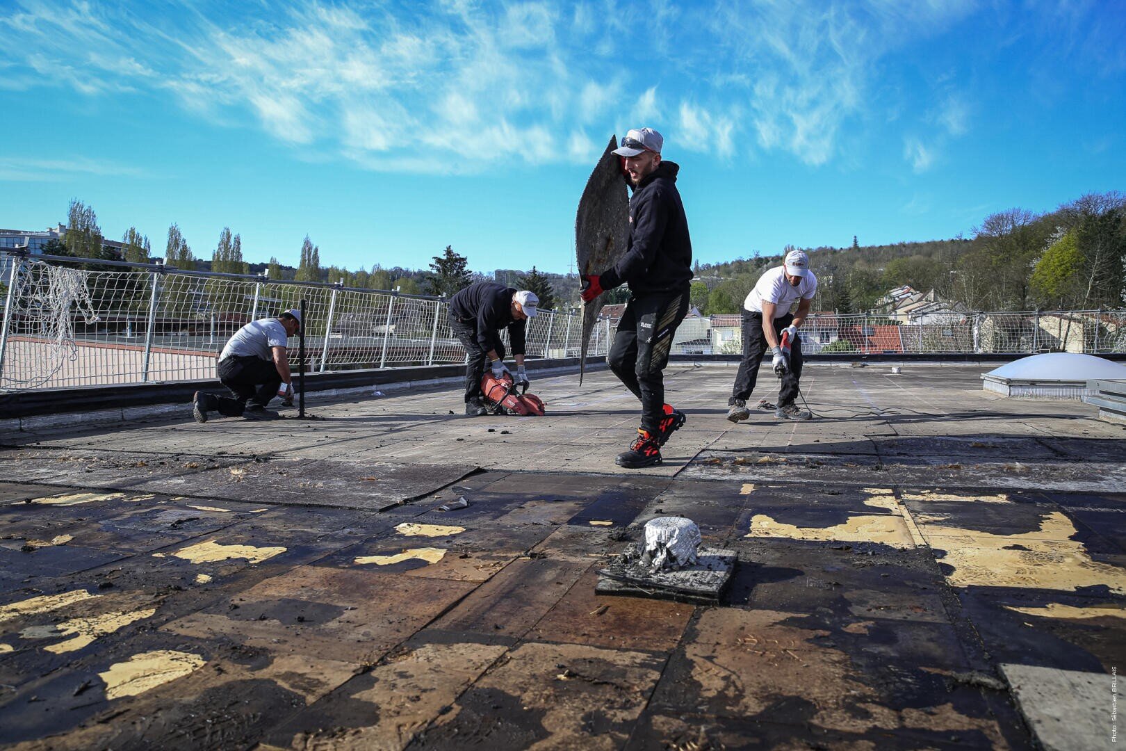 Vier Arbeiter mit Mützen und Handschuhen entfernen an einem klaren, sonnigen Tag altes Dachmaterial von einem Flachdach. Ein Arbeiter im Vordergrund trägt ein großes Stück Schutt, während die anderen das Material mit Werkzeugen anheben.