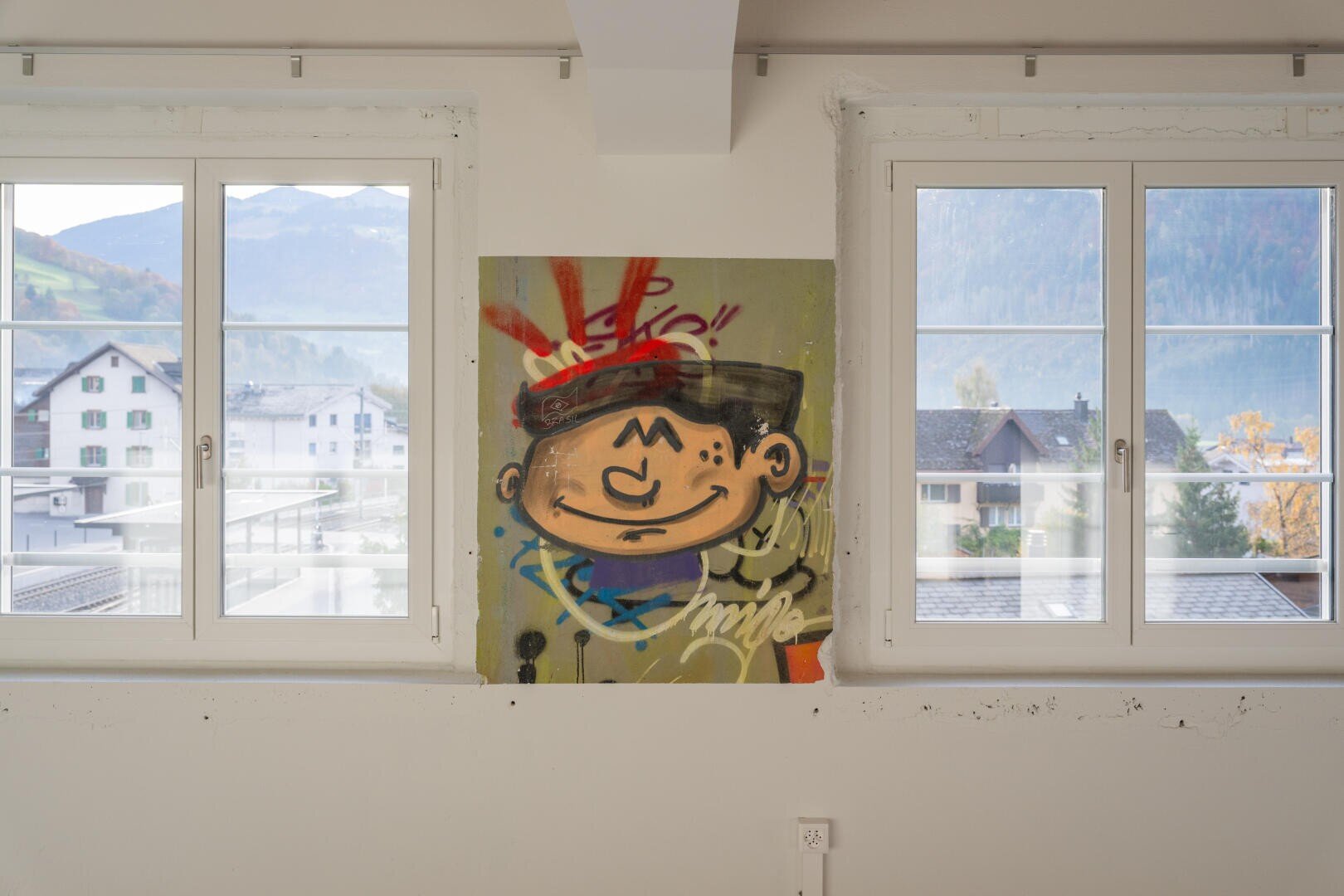Ein farbenfrohes Graffiti im Cartoon-Stil mit einem lächelnden Gesicht ist auf eine Leinwand gemalt, die zwischen zwei weiß gerahmten Fenstern platziert ist, aus denen Berge und Häuser zu sehen sind.