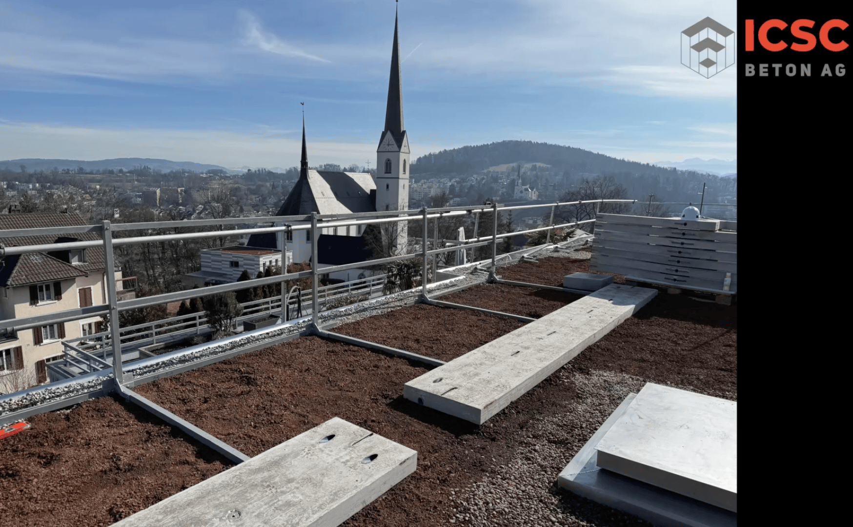 Un chantier de construction sur un toit avec des balustrades métalliques, des plaques de béton et de la terre. En arrière-plan, on peut voir des maisons, des arbres, des collines et une église avec deux hauts clochers. Dans le coin supérieur droit, on peut voir le logo d'ICSC Beton AG.