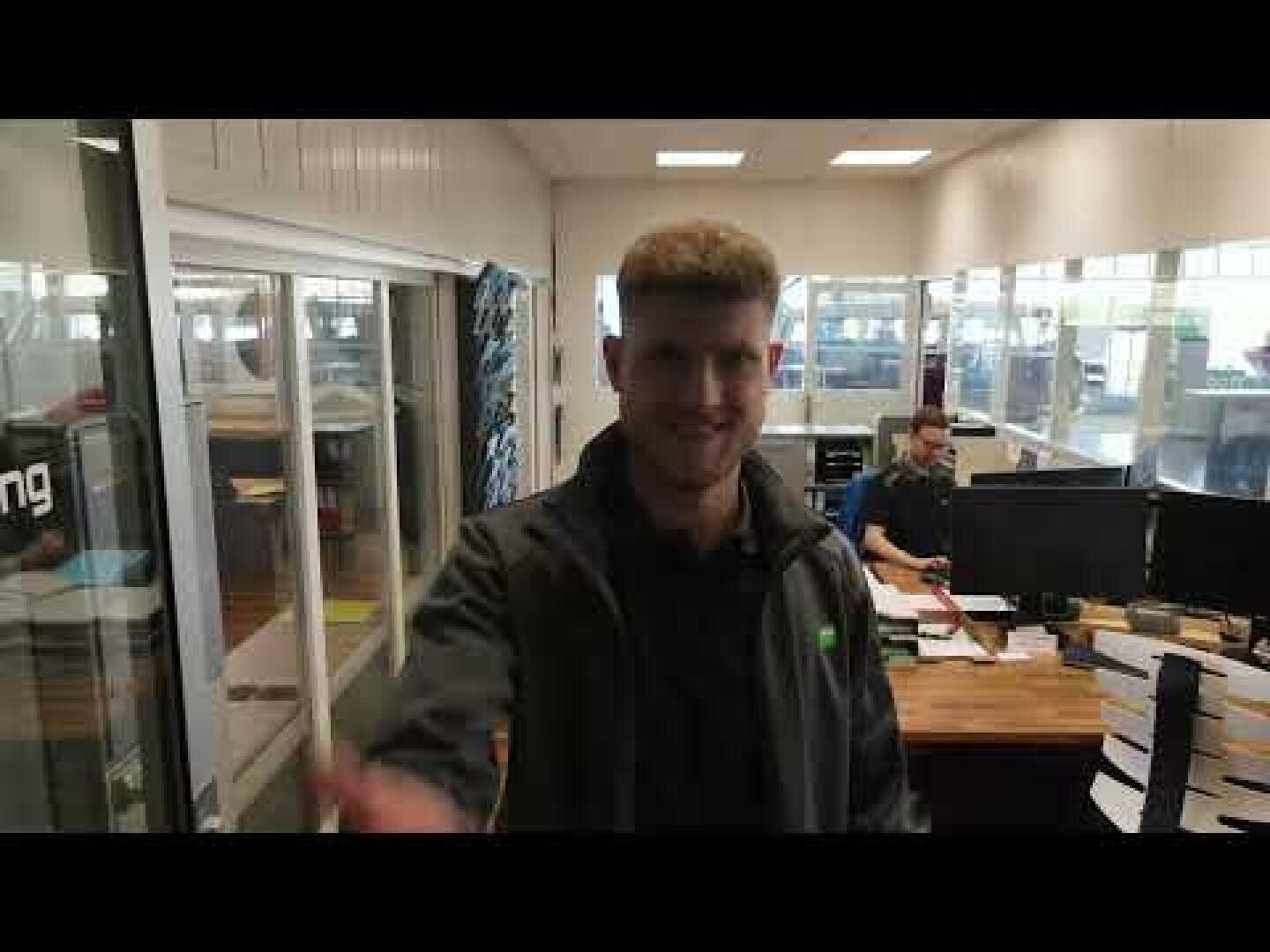 Ein Mann steht lächelnd im Vordergrund eines Büroraums, in dem Schreibtische, Papiere und Computer zu sehen sind. Eine andere Person arbeitet an einem Schreibtisch im Hintergrund in der Nähe großer Fenster.