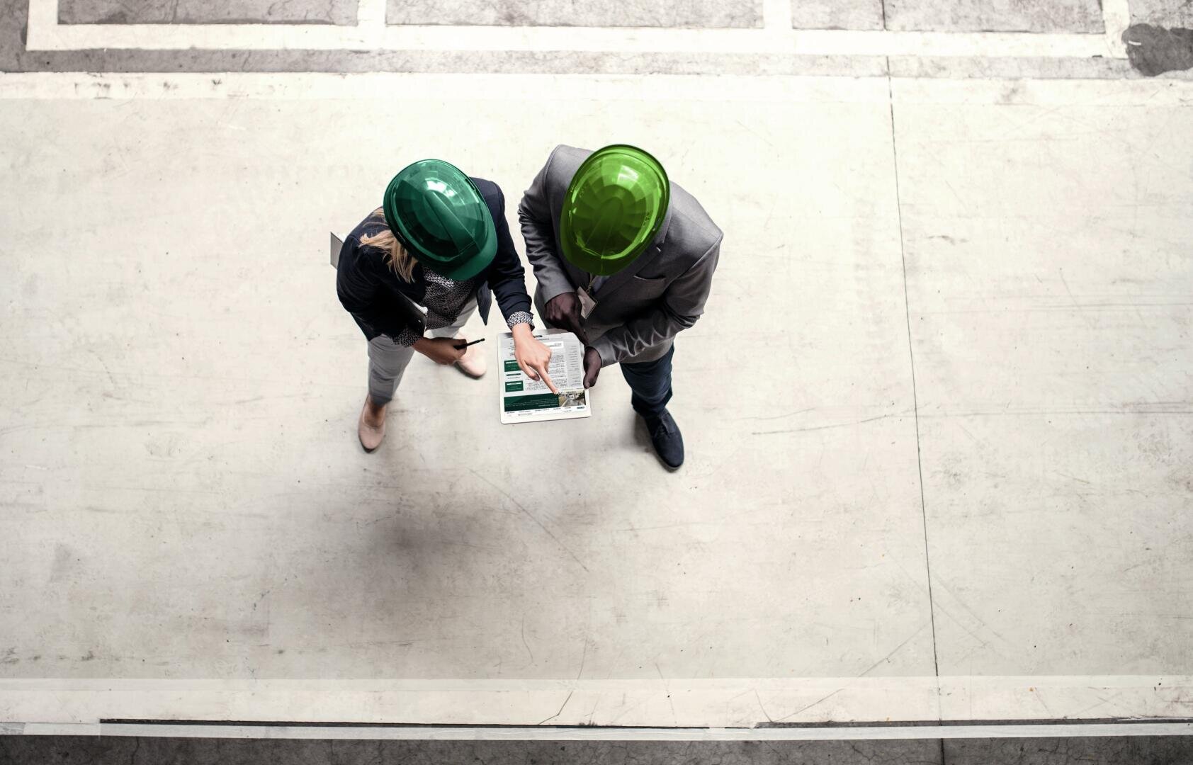 Zwei Personen mit grünen Schutzhelmen stehen auf einem Betonboden und betrachten und besprechen Dokumente oder Blaupausen auf einem Klemmbrett, das von oben betrachtet wird.