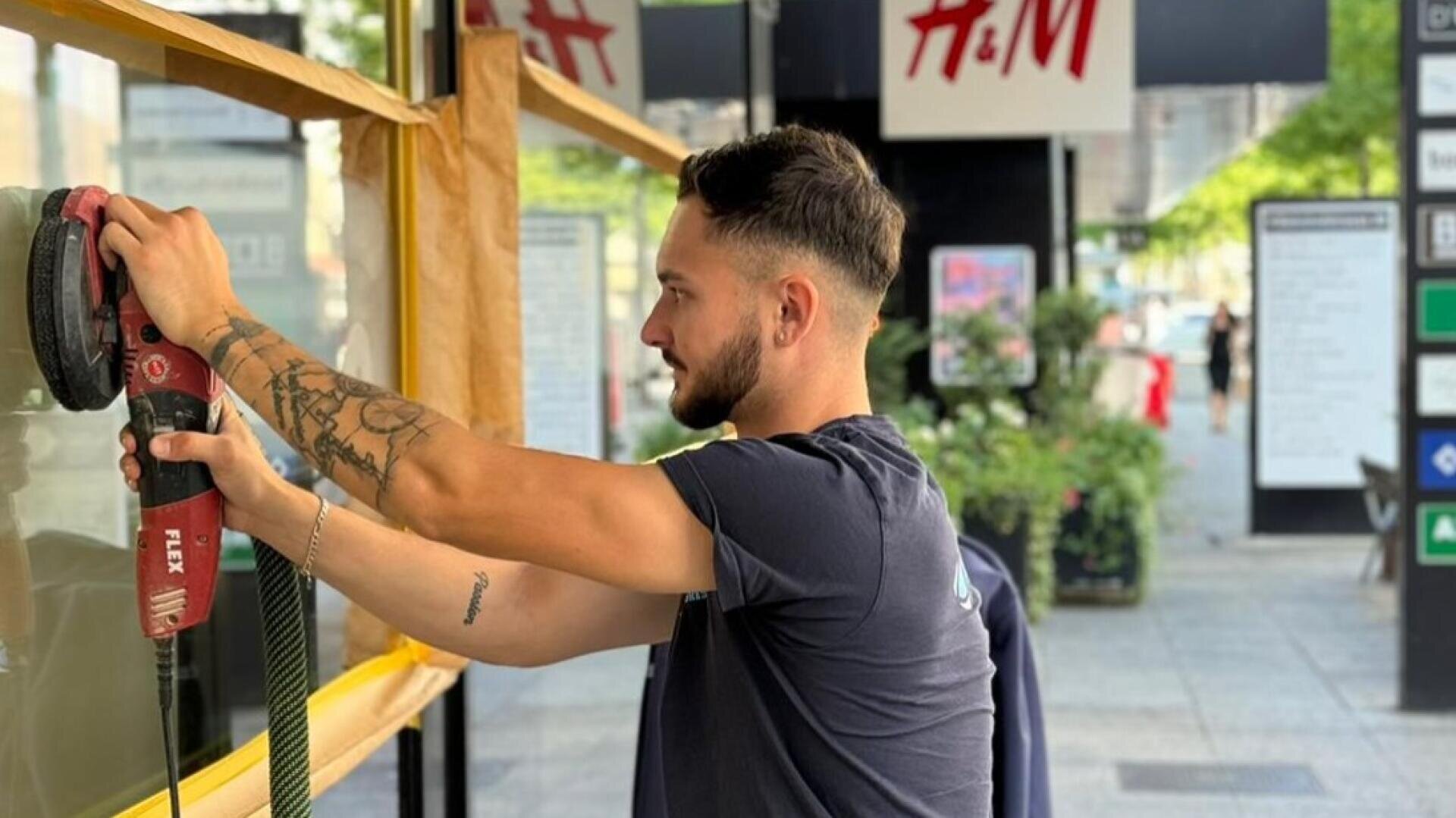 Ein Mann mit Tattoos benutzt ein Elektrowerkzeug, um ein Fenster in einer Straße in der Stadt zu polieren oder zu reinigen, während im Hintergrund H&M-Schilder und Grünflächen zu sehen sind.
