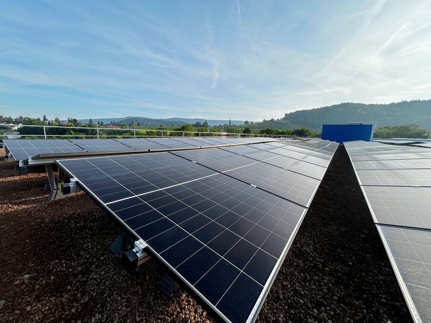 Reihen von Solarmodulen sind auf einem Dach unter strahlend blauem Himmel installiert, und im Hintergrund sind grüne Hügel zu sehen.