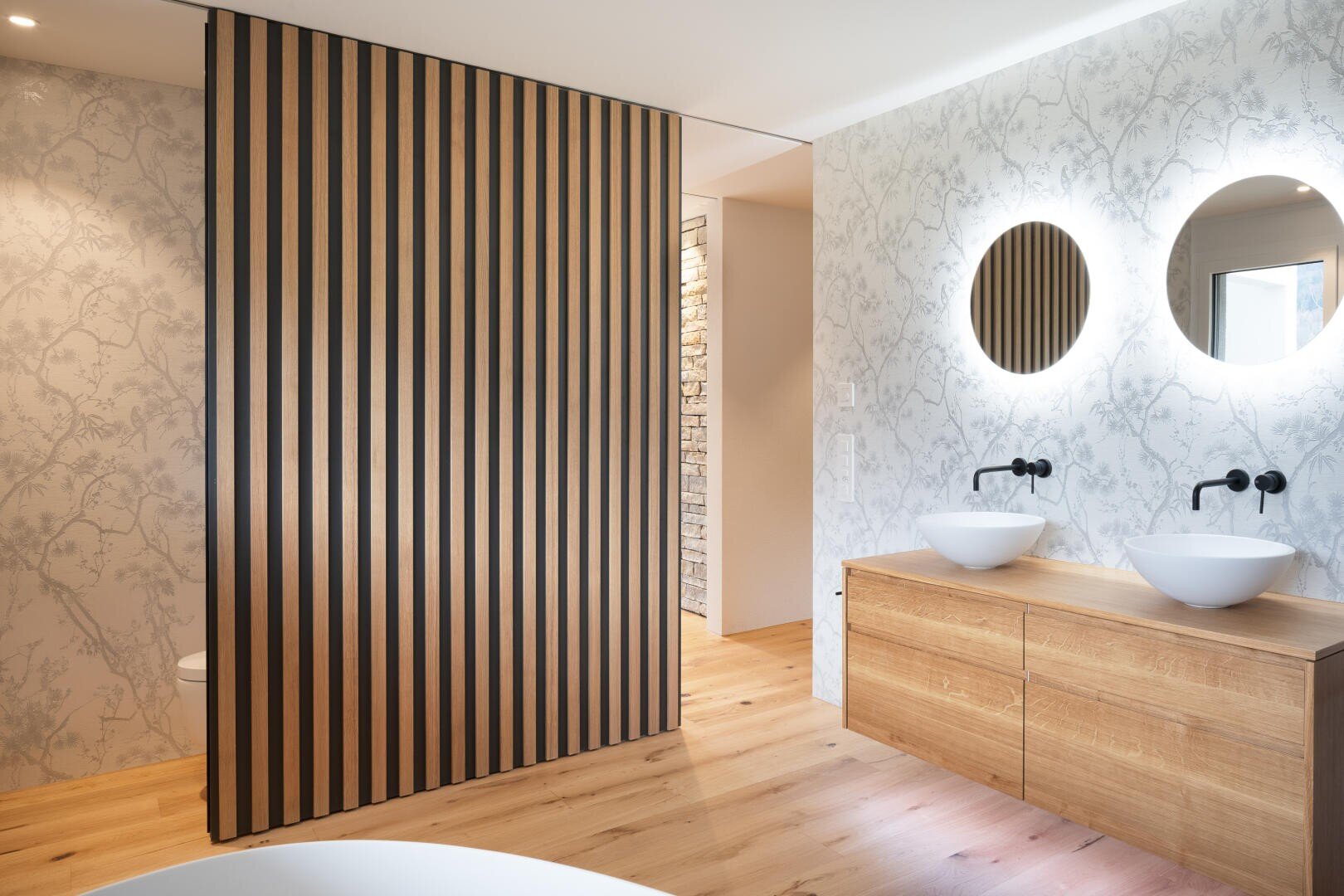 Modernes Badezimmer mit hellem Holzfußboden, einem hölzernen Waschtisch mit zwei Gefäßwaschbecken und runden Spiegeln, die von hinten beleuchtet sind und ein sanftes Licht verbreiten. Eine Trennwand aus Lattenholz schirmt den Toilettenbereich teilweise ab. Eine dezent gemusterte Tapete ziert die Wände.