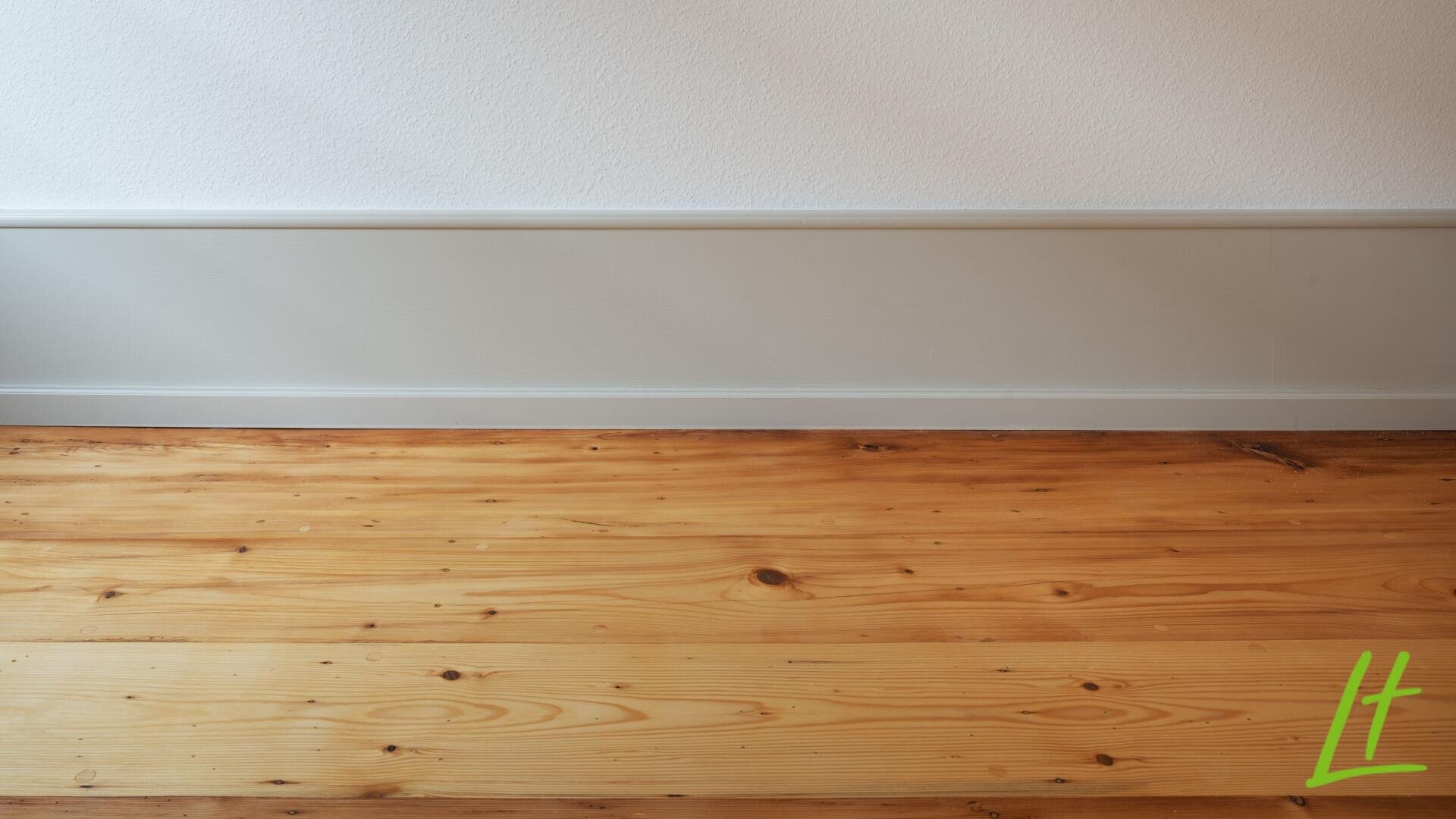 Ein heller Holzdielenboden trifft auf eine weiße Wand mit einer hellgrauen Fußleiste; in der rechten unteren Ecke befindet sich ein grünes L-Logo.