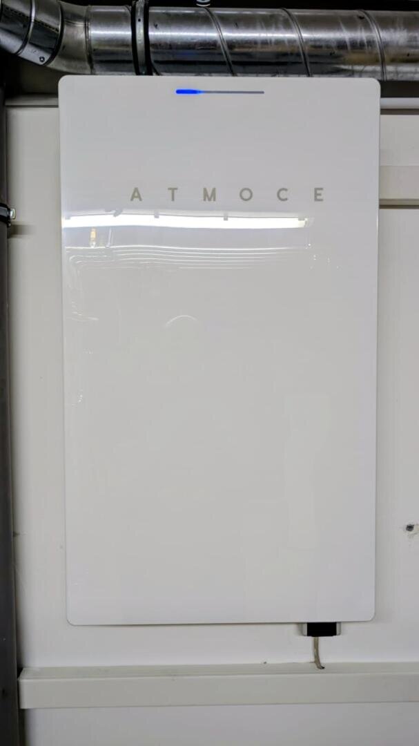 Ein großes, weißes, rechteckiges Gerät mit der Aufschrift ATMOC ist an einer Wand angebracht. Oben ist ein kleines blaues Licht zu sehen, und unten ist ein Kabel angeschlossen. An der Decke über dem Gerät befinden sich Leitungen und Rohre.