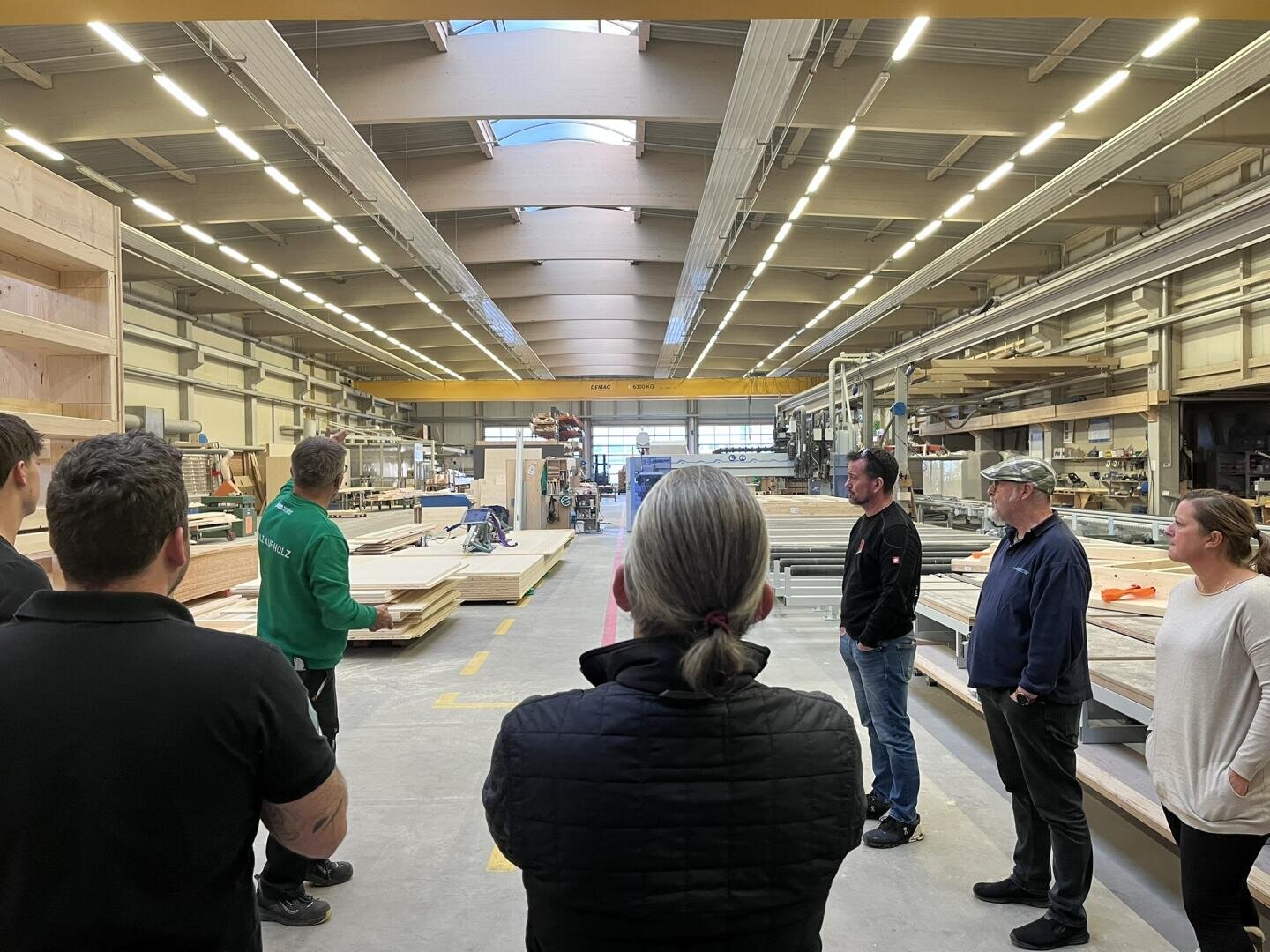 Eine Gruppe von Personen steht in einer geräumigen, gut beleuchteten Holzverarbeitungsfabrik und hört einer Person in einer grünen Jacke zu, die sie anscheinend anleitet oder instruiert. Im Hintergrund sind Holzstapel und Maschinen zu sehen.