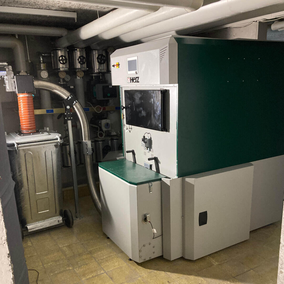 Eine große Industriemaschine mit grünen und weißen Platten und der Aufschrift HERZ steht in einem Technikraum mit Rohren an der Decke und Steuerventilen an der Wand. Der Boden ist gefliest, und der Raum wirkt sauber und ordentlich.