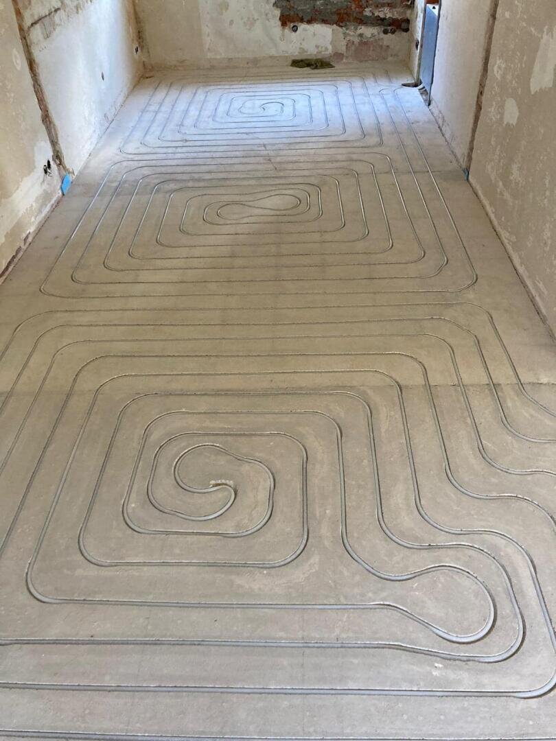 Metallrohre sind in einem spiralförmigen und rechteckigen Muster auf einem nackten Betonboden verlegt, wahrscheinlich für die Installation einer Fußbodenheizung in einem zu renovierenden Raum. Die Wände scheinen unfertig zu sein.