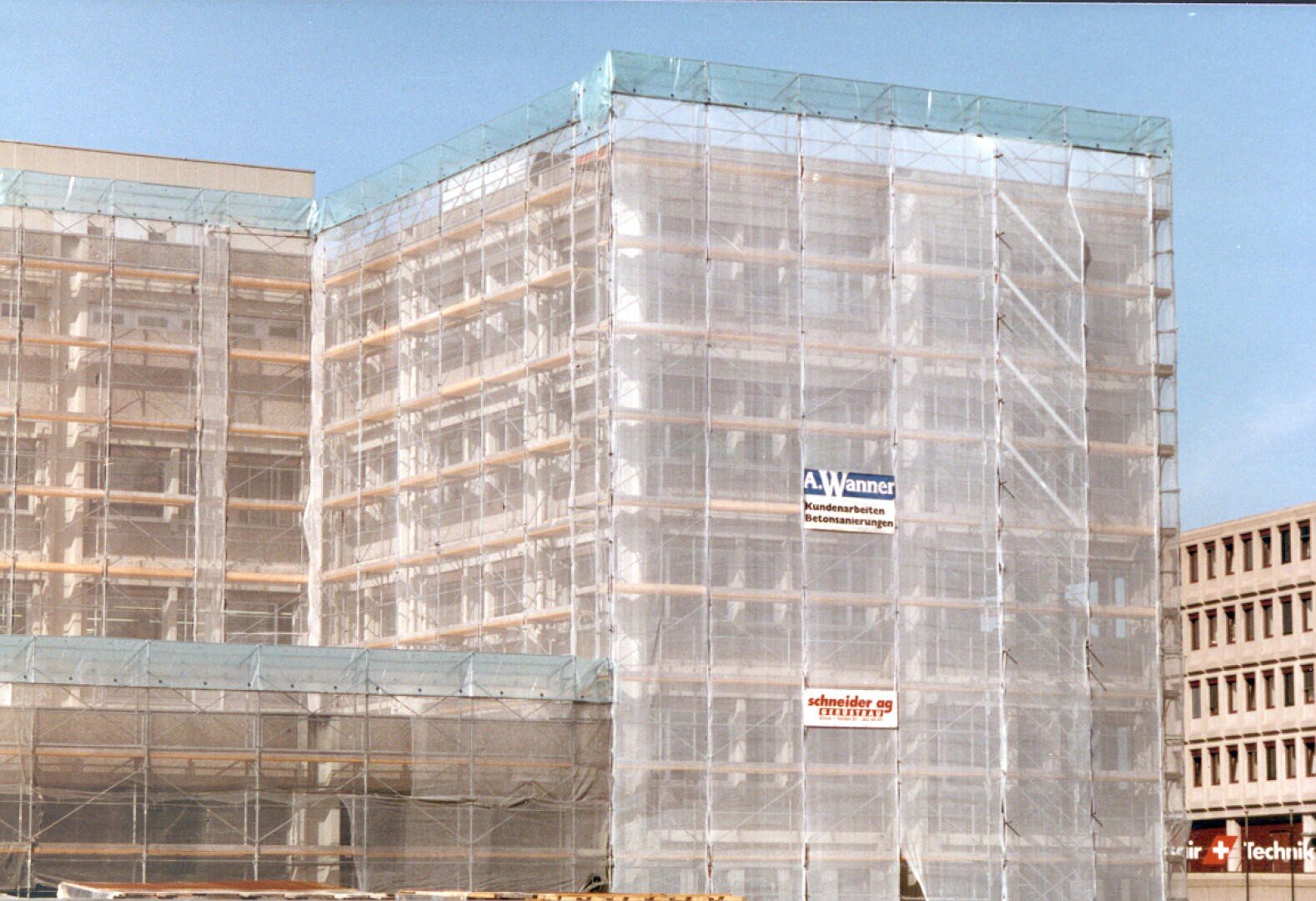 Ein großes Gebäude mit Gerüsten und Schutznetzen, an denen Firmenschilder befestigt sind, vor einem strahlend blauen Himmel.