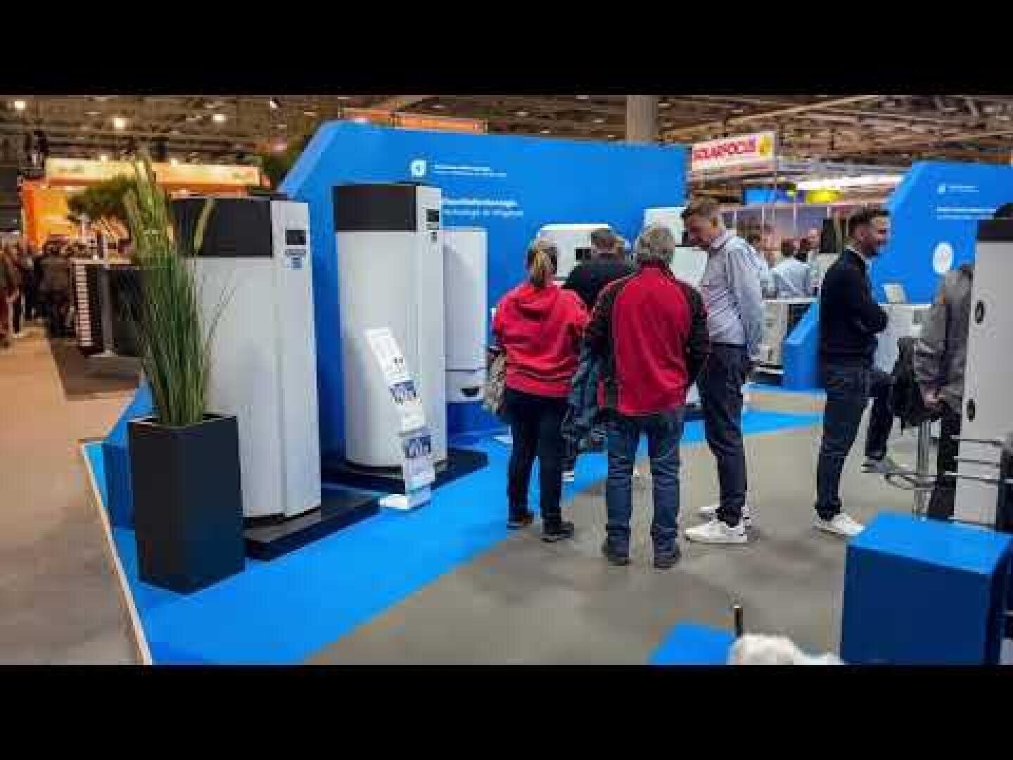 Eine Gruppe von Menschen steht vor großen weißen Geräten an einem blauen Messestand und beschäftigt sich mit Ausstellungsmaterial und miteinander. Im Hintergrund sind andere Stände und Besucher zu sehen.