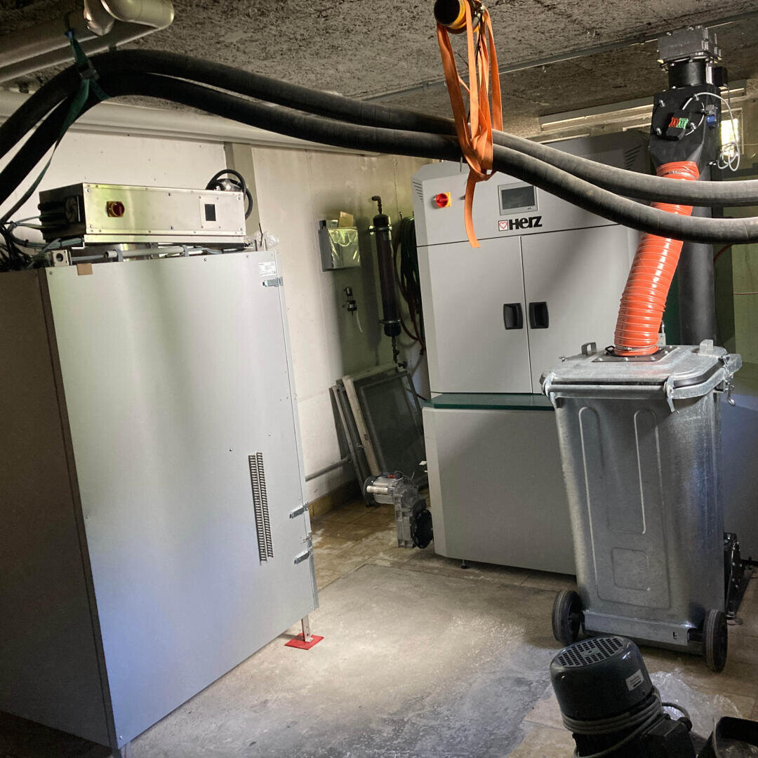 Ein Kellerraum mit industriellen Geräten, darunter große Metallschränke, dicke schwarze und orangefarbene Schläuche, ein Abfalleimer und verschiedene an den Wänden montierte Stromkästen. Der Raum wirkt funktional und utilitaristisch.