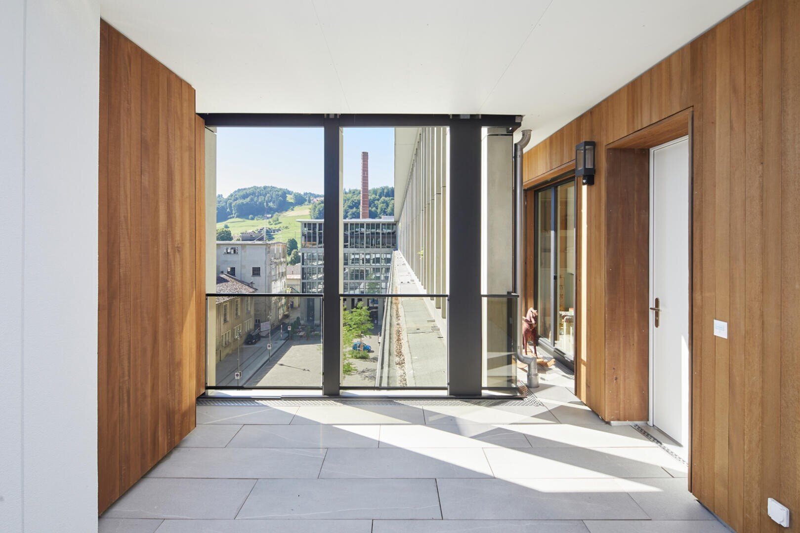 Un balcon moderne avec des murs en bois, de grandes baies vitrées et des carreaux de sol clairs offre une vue sur une rue et des collines vertes lointaines sous un ciel dégagé. La lumière du soleil y pénètre et projette des ombres sur le sol.