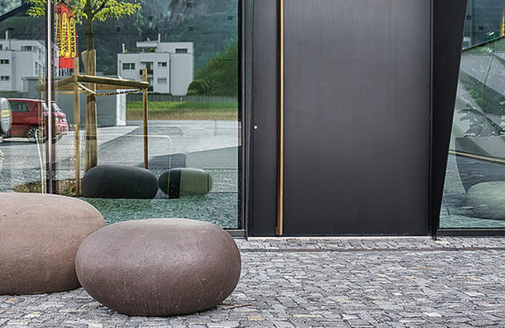 Zwei große, runde Steinbänke stehen auf einem Kopfsteinpflaster vor einem modernen Gebäude mit großen reflektierenden Glasfenstern. In den Fenstern spiegeln sich nahe gelegene Gebäude, ein Baum und ein rotes Auto.