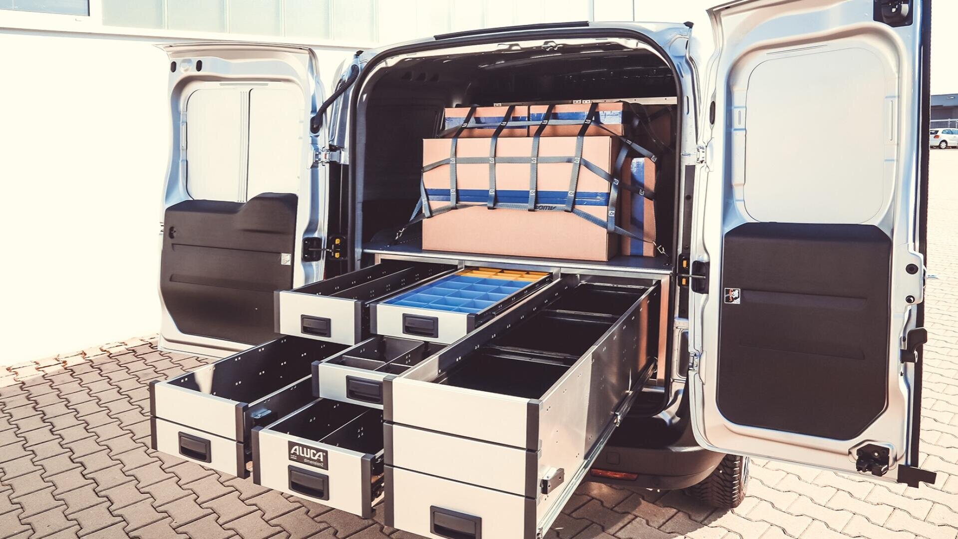 Ein Lieferwagen mit geöffneten Hecktüren zeigt einen organisierten Stauraum: Mehrere Metallschubladen sind herausgezogen, und im Inneren des Fahrzeugs sind mit Gurten gesicherte Kisten gestapelt.