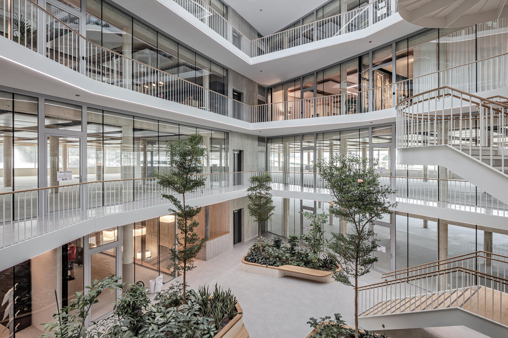 Ein modernes, mehrstöckiges Büro-Atrium mit großen Glasfenstern, weißen Geländern, mehreren Zimmerbäumen, Pflanzen und Holzbänken. Offene Gänge und Treppenhäuser schaffen eine helle, großzügige Atmosphäre.