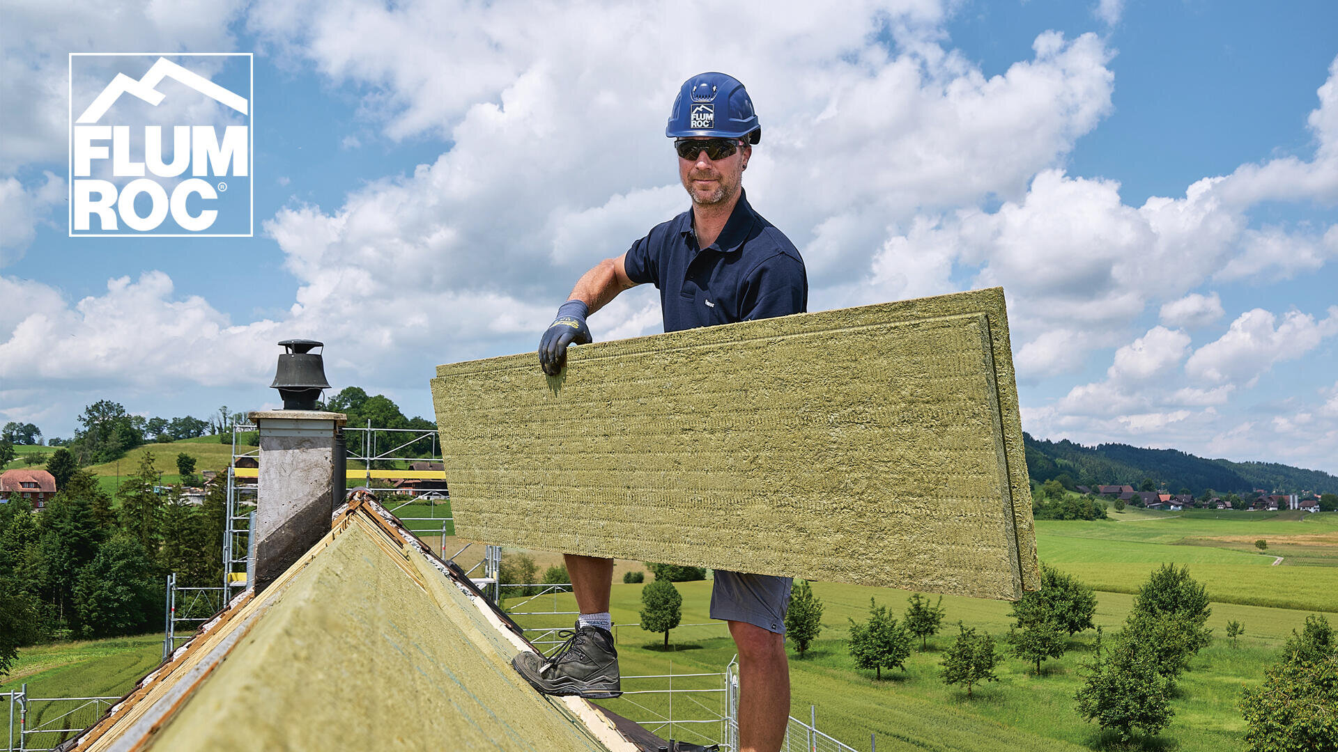 Ein Bauarbeiter in Schutzkleidung steht auf einem Dach und hält ein großes Stück Dämmmaterial in der Hand. Die Szene bietet einen Blick auf grüne Felder unter einem blauen Himmel. In der oberen linken Ecke ist ein FLUMROC-Logo zu sehen.