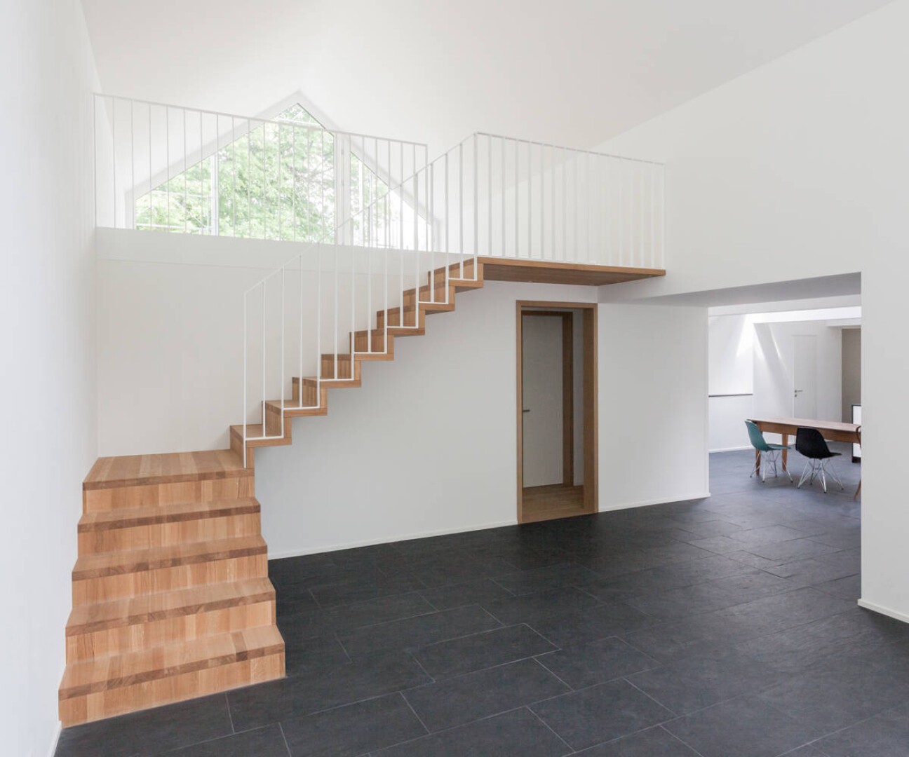 Minimalistisches Interieur mit Holztreppe und weißem Geländer, das in eine obere Etage führt, schwarzem Fliesenboden, weißen Wänden und einem Tisch mit Stühlen im angrenzenden Raum. Großes Fenster sorgt für Tageslicht.