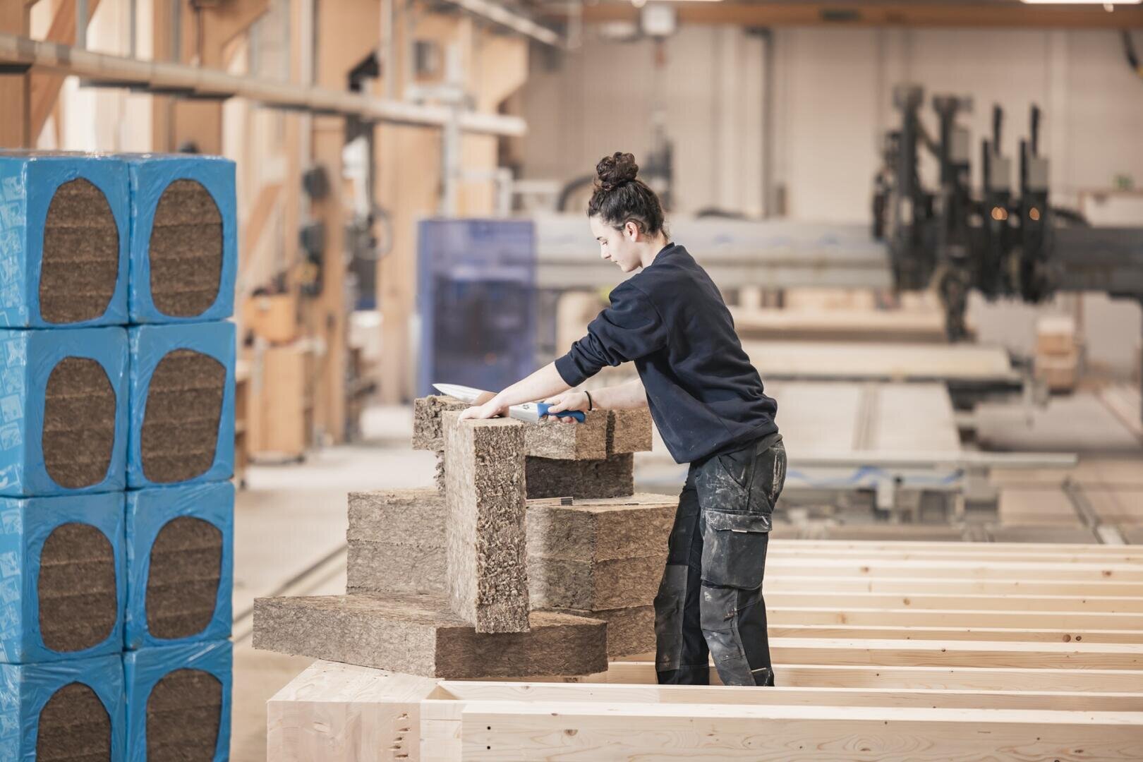 Eine Person in Arbeitskleidung stapelt in einer geräumigen, gut beleuchteten Werkstatt braune Dämmplatten auf Holzbalken. Auf der linken Seite sind blaue Isolierblöcke zu sehen.
