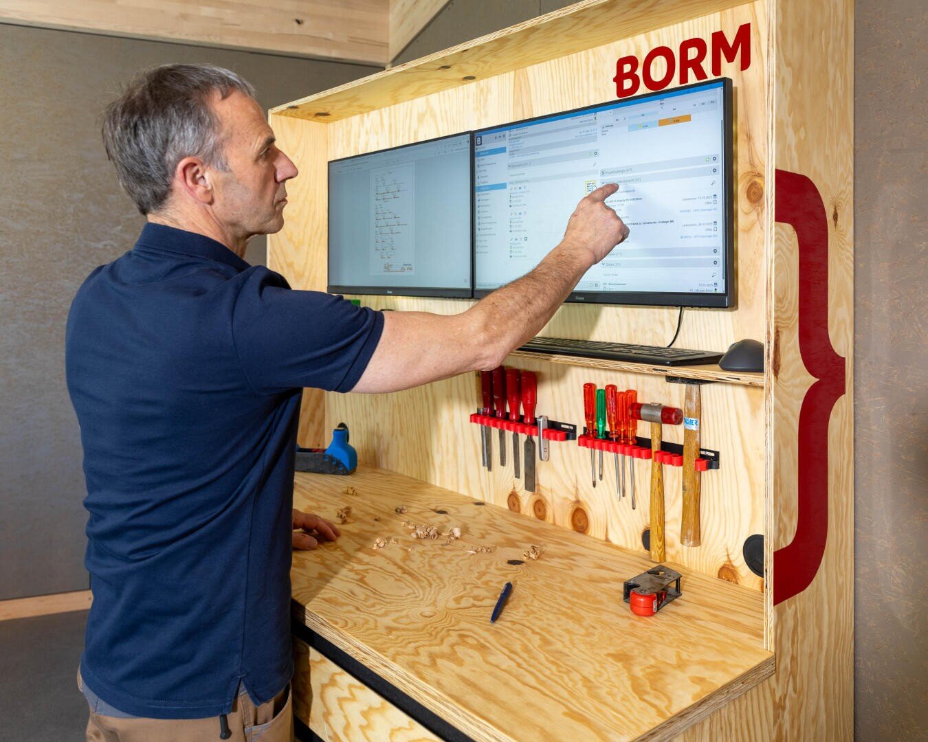 Ein Mann in einem marineblauen Hemd bedient einen Touchscreen-Monitor, der auf einem hölzernen Arbeitsplatz mit Werkzeugen, Holzspänen und einem Maßband montiert ist; über dem Bildschirm wird das Wort BORM angezeigt.