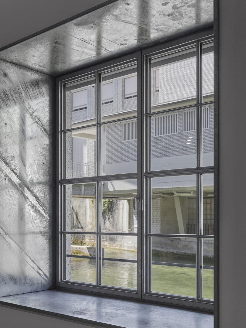 Ein großes Fenster mit Metallrahmen bietet einen Blick auf einen Kanal und ein modernes Gebäude mit Ziegel- und Glaselementen. Das Sonnenlicht wirft Schatten auf das Fensterbrett und die Innenwand.