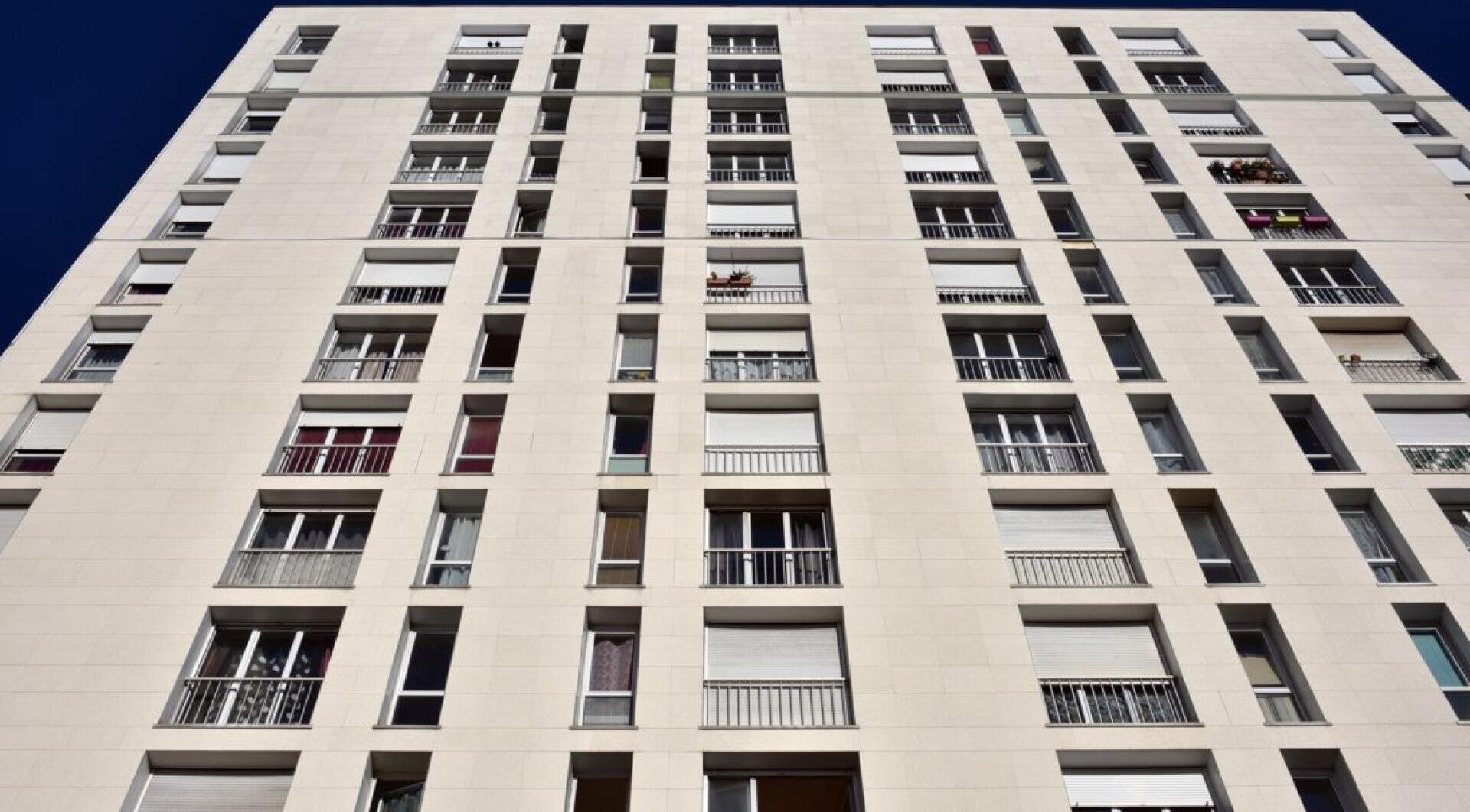 Ein hohes, modernes Wohngebäude mit weißer Fassade, vielen Fenstern und mehreren Balkonen, auf denen Pflanzen oder Gegenstände stehen. Das Foto ist von unten aufgenommen, mit Blick auf das Gebäude vor einem klaren blauen Himmel.