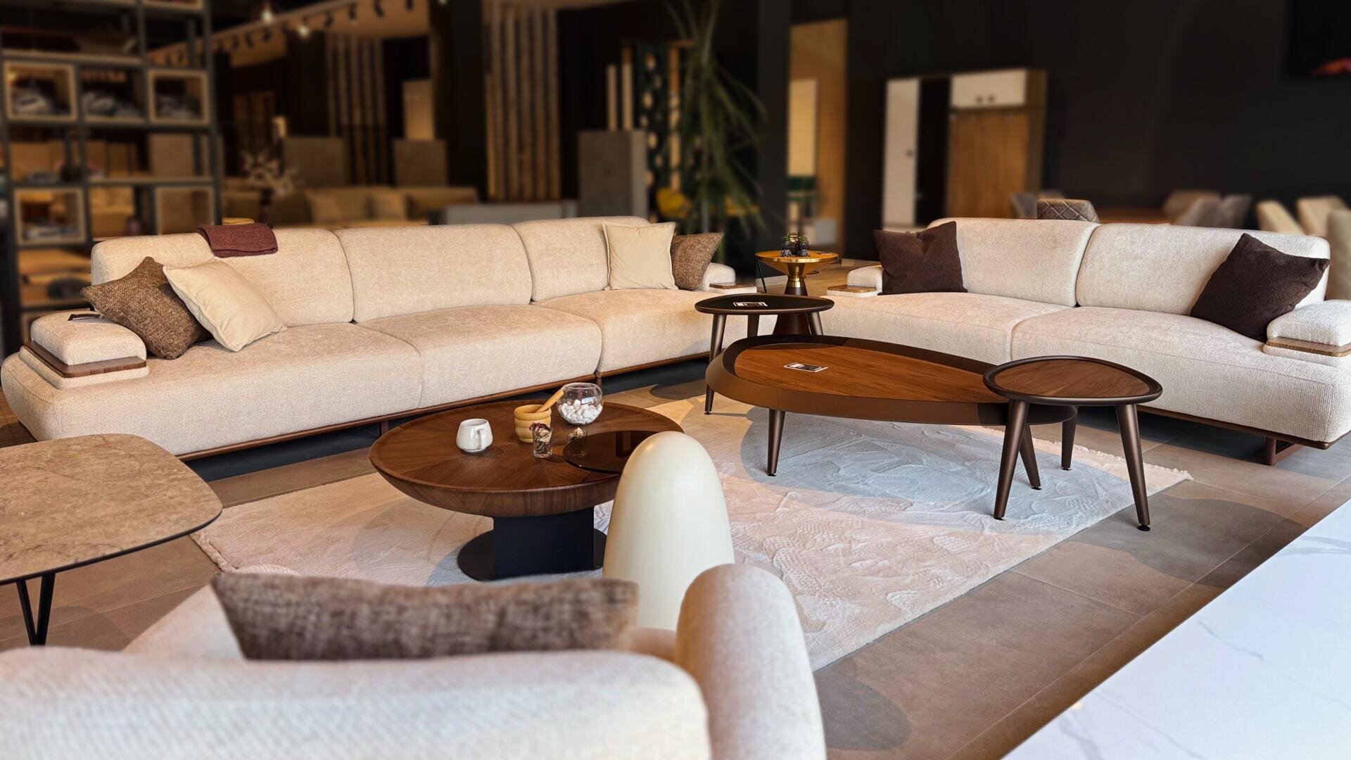 Ein modernes Wohnzimmer mit beigen Sofas, braunen Akzentkissen und hölzernen Couchtischen auf einem hellen Teppich, umgeben von sanfter Beleuchtung und zeitgenössischem Dekor.