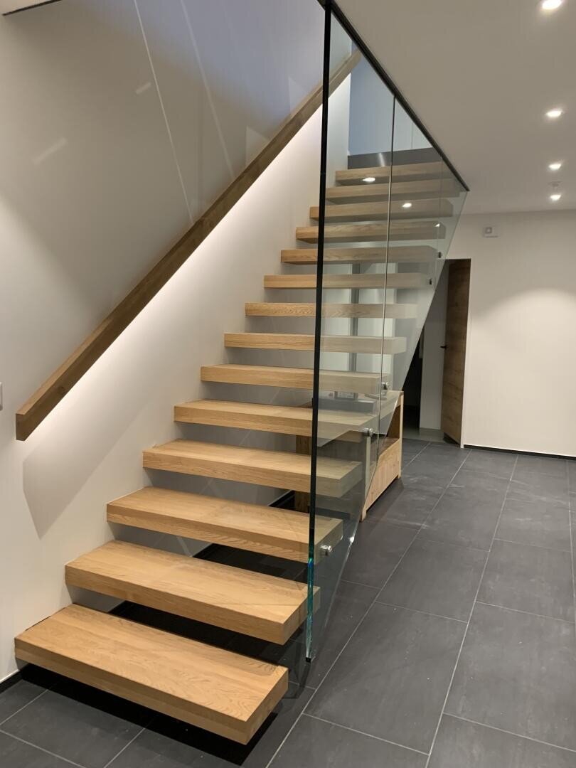 Un escalier flottant moderne en bois avec une rampe en verre dans un intérieur minimaliste, avec des carreaux de sol gris foncé et des murs blancs avec des plafonniers encastrés.