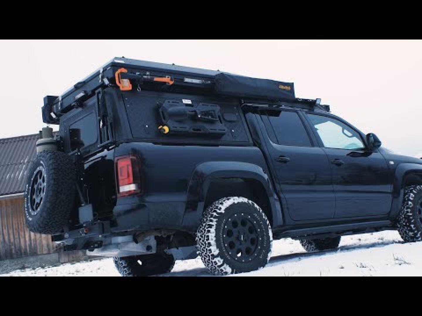 Ein schwarzer, geländegängiger Pickup mit großen Reifen ist mit Camping- und Überlebensausrüstung auf der Ladefläche ausgestattet und parkt im Schnee neben einem Holzgebäude. Auf dem Lkw sind Staukästen und eine Markise montiert.
