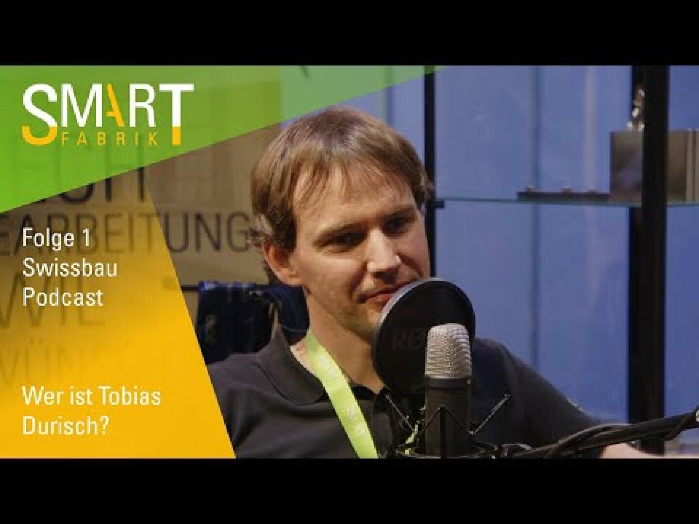 Ein Mann mit braunem Haar spricht während einer Podcast-Aufnahme in ein Mikrofon. Der Text auf dem Bild lautet: SMART FABRIK, Folge 1 Swissbau Podcast, Wer ist Tobias Durisch? Grüne und gelbe Grafiken befinden sich in der oberen linken Ecke.