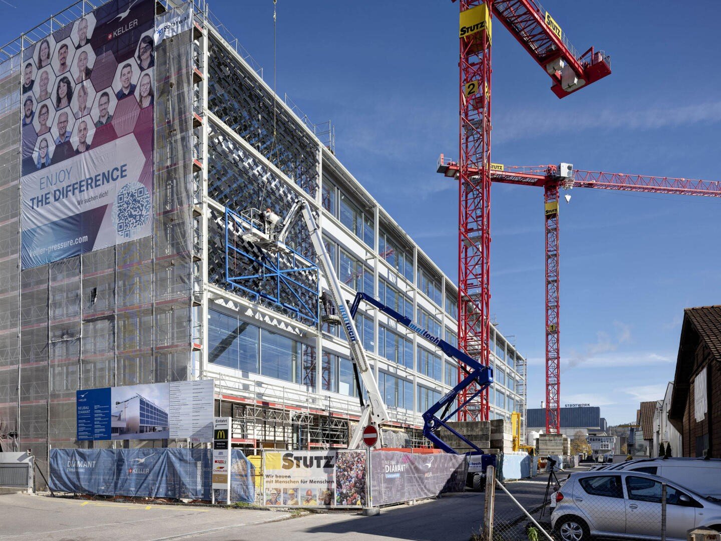 Un bâtiment moderne en construction, recouvert d'échafaudages et de bannières. Deux grues et un ascenseur de chantier sont visibles, et des ouvriers sont présents. Plusieurs voitures sont garées à proximité et le ciel est bleu.