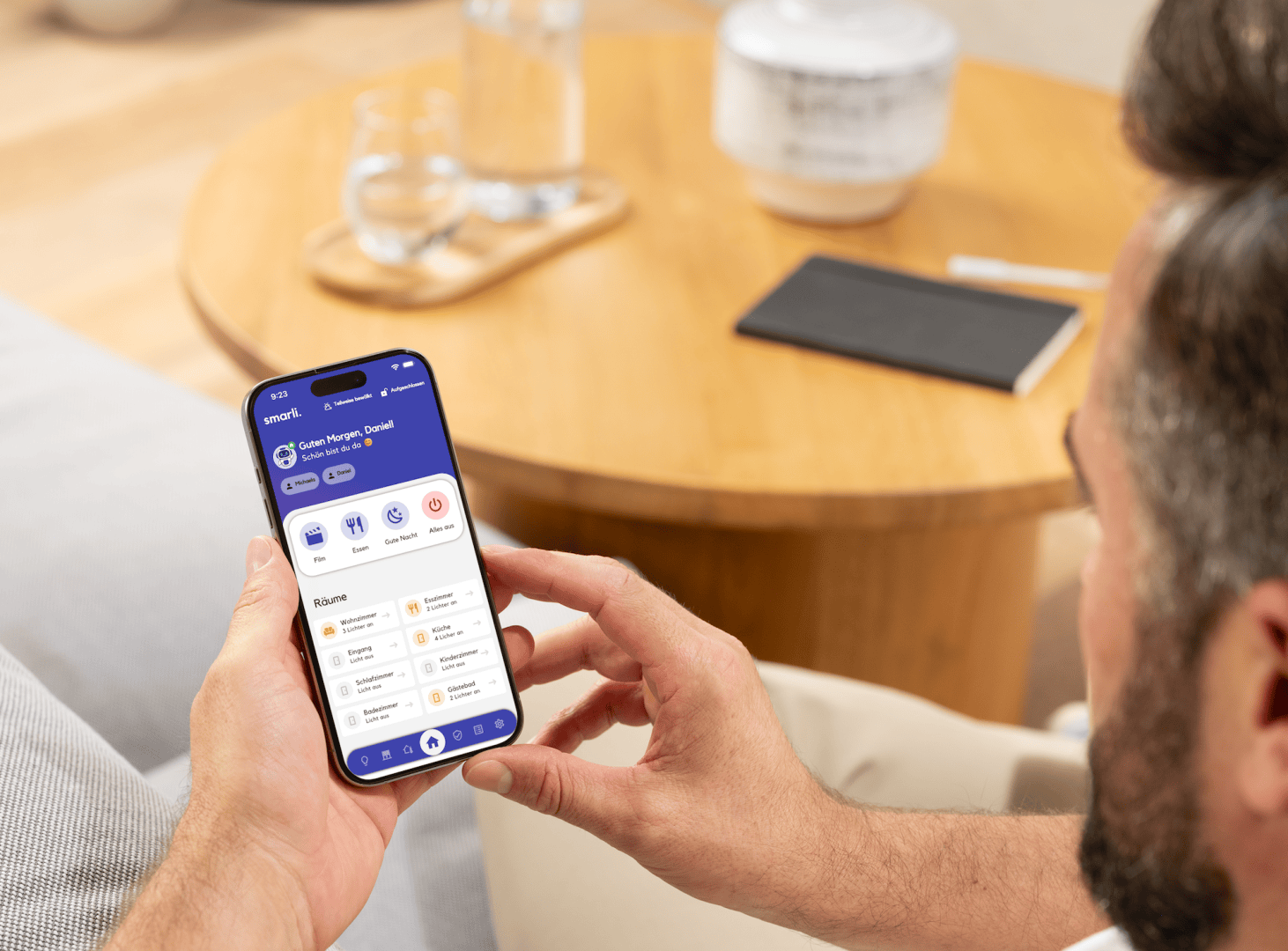 Eine Person sitzt auf einer Couch und hält ein Smartphone in der Hand, auf dem eine Gesundheits- und Fitness-App mit Optionen für Schritte, Kalorien, Training und Herzfrequenz angezeigt wird. Im Hintergrund steht ein hölzerner Couchtisch mit einem Notizbuch und einem Glas.