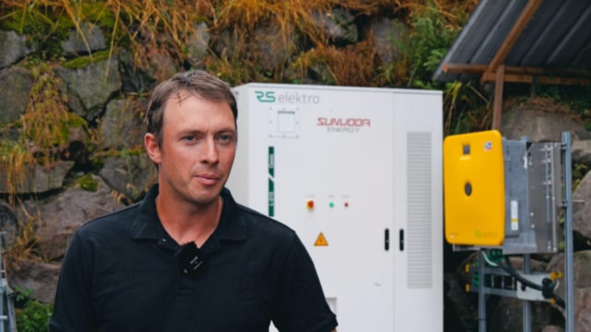 Ein Mann in einem schwarzen Hemd steht im Freien vor einer elektrischen Anlage und einer Felswand, im Hintergrund sind einige Pflanzen und Gras zu sehen.