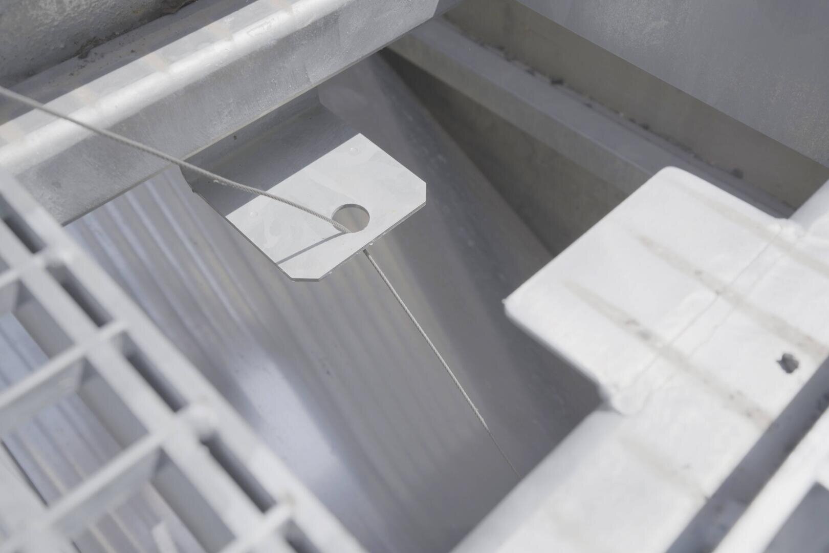 Gros plan sur une structure métallique industrielle avec une plateforme grillagée, une surface striée et une plaque métallique avec un trou rond reliée par un fil ou un câble tendu.