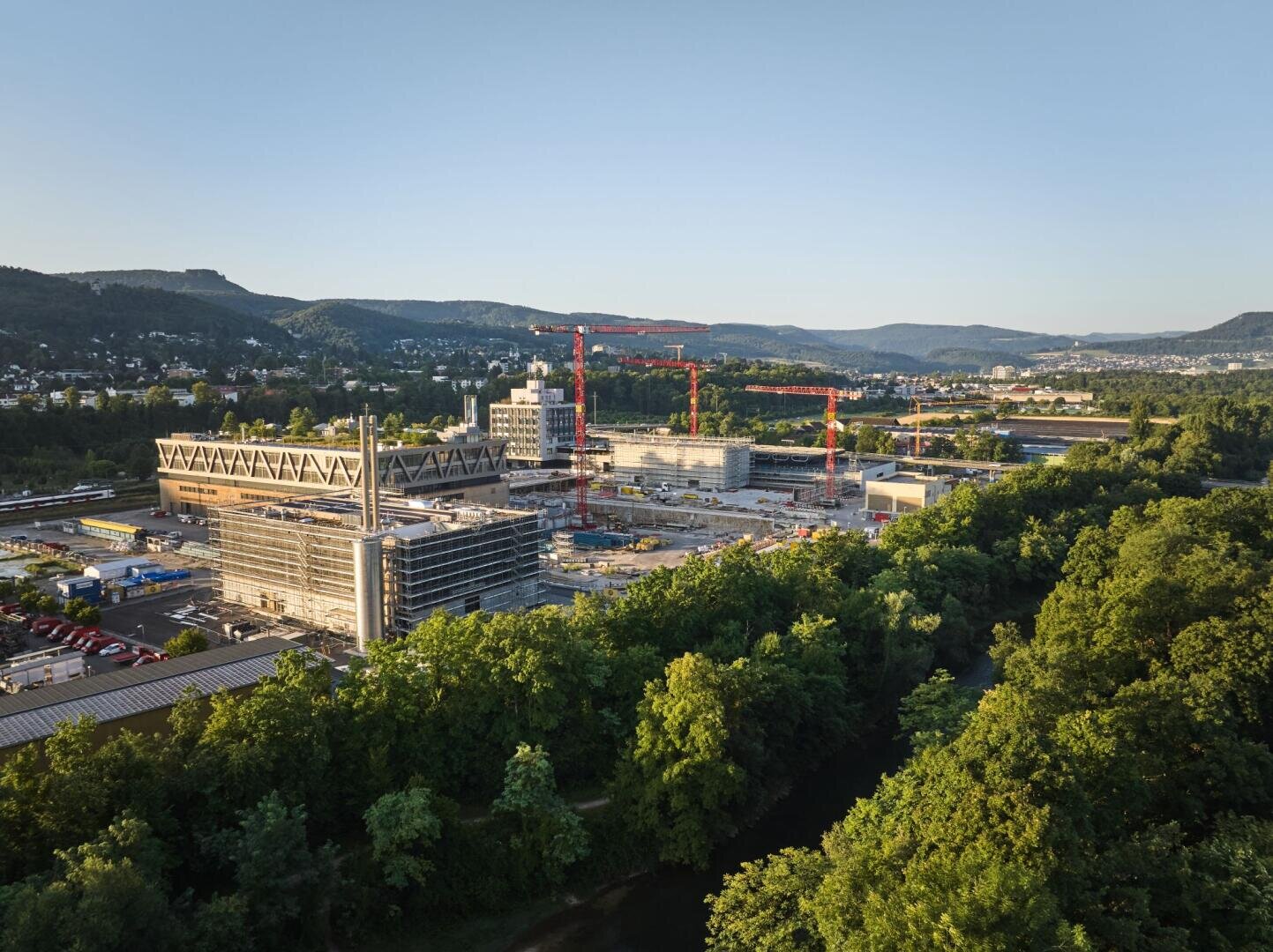 Luftaufnahme einer Großbaustelle mit mehreren Kränen, teilweise fertiggestellten modernen Gebäuden und umliegenden Bäumen; unter klarem Himmel sind in der Ferne Hügel und die Stadtlandschaft zu sehen.