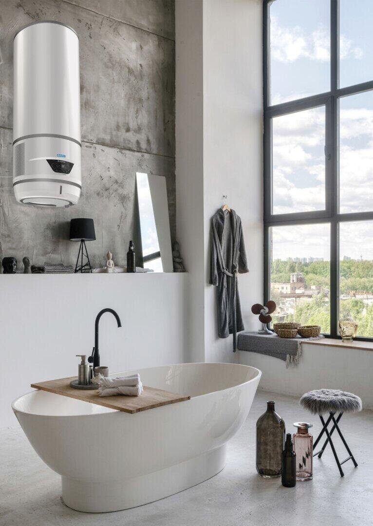 Modernes Badezimmer mit einer freistehenden Badewanne, einem an der Wand montierten Warmwasserbereiter, großen Fenstern und natürlichem Licht. Zur Ausstattung gehören ein schwarzer Wasserhahn, ein Bademantel, ein Hocker sowie verschiedene Flaschen und Accessoires auf dem Boden und der Wannenablage.