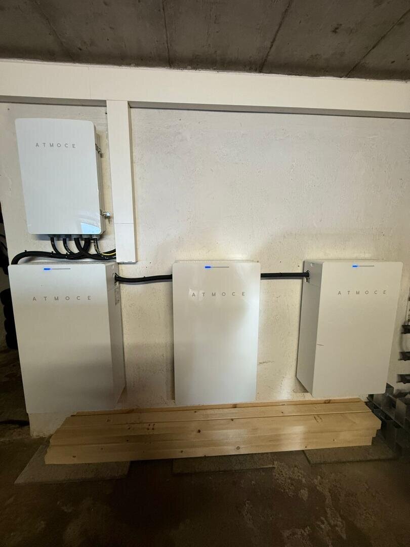 Drei große weiße elektrische Batteriespeicher mit der Aufschrift ATMOCE sind an einer weißen Wand angebracht und mit schwarzen Kabeln verbunden. Der Bereich scheint ein Versorgungs- oder Lagerraum zu sein.