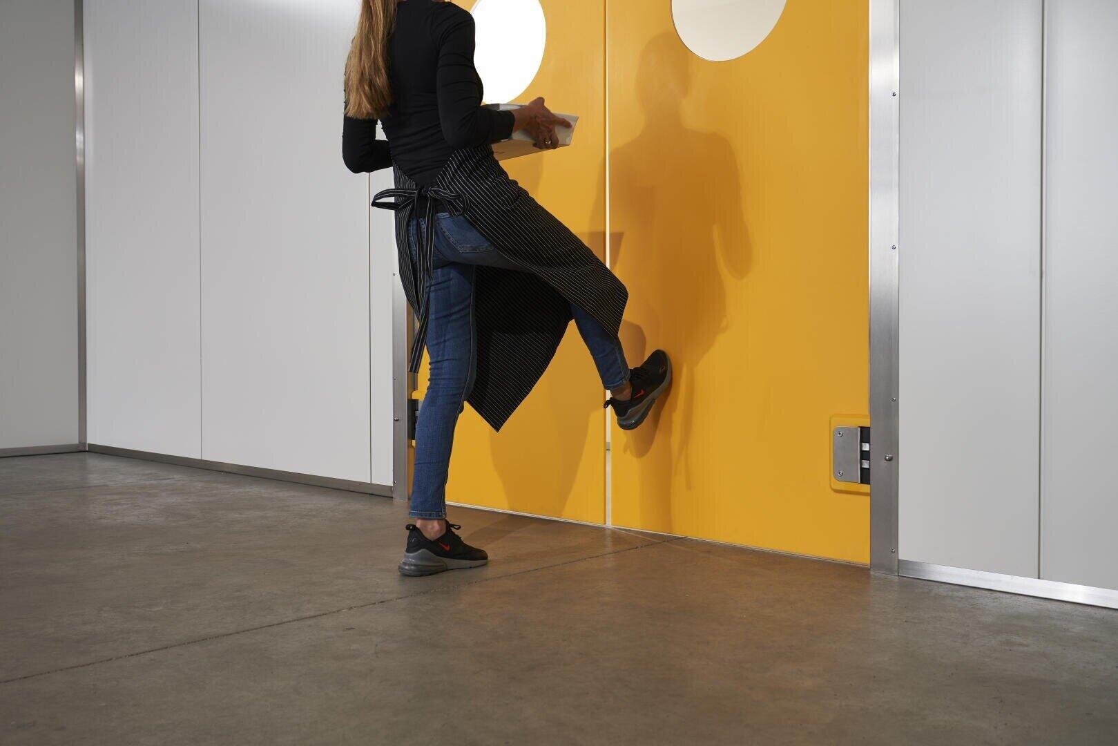 Eine Person, die eine Schürze und Jeans trägt, öffnet eine leuchtend gelbe Tür mit einem Fußpedal, während sie Gegenstände in ihren Händen hält.
