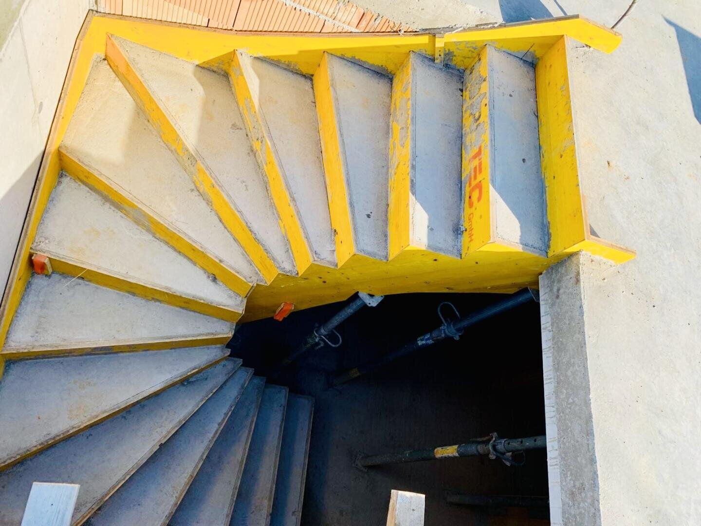Eine im Bau befindliche Wendeltreppe aus Beton mit gelber Sicherheitsumrandung und darunter sichtbarem Metallgerüst. Helles Sonnenlicht wirft starke Schatten auf die Treppe und die umliegenden Flächen.