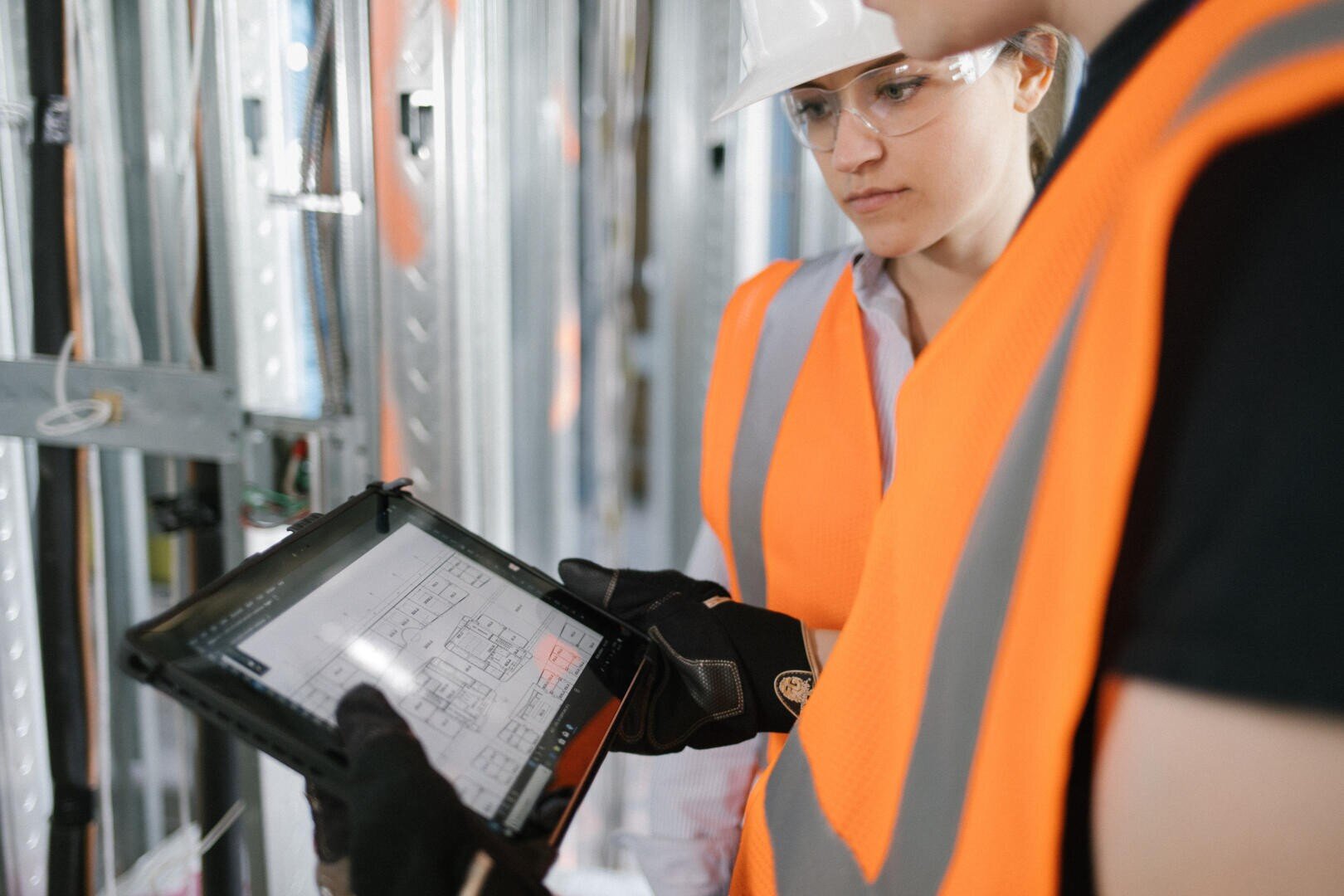 Zwei Bauarbeiter in orangefarbenen Sicherheitswesten und Schutzhelmen schauen auf ein digitales Tablet, das Baupläne in einem teilweise errichteten Gebäude mit Metallgerüst anzeigt.
