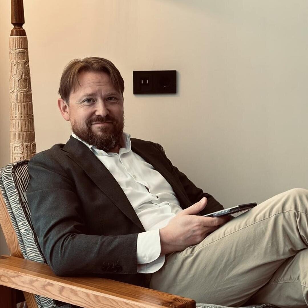 Ein Mann mit Bart, der einen dunklen Blazer, ein weißes Hemd und eine beigefarbene Hose trägt, sitzt auf einem Holzstuhl, hält ein Smartphone und lächelt in die Kamera. An der Wand hinter ihm befinden sich ein geschnitzter Holzpfosten und eine Steckdose.