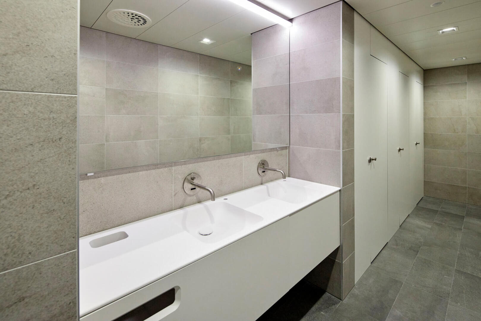 Moderne öffentliche Toilette mit einem langen weißen Waschbecken mit zwei Wasserhähnen, einem großen Wandspiegel und mehreren geschlossenen Kabinentüren mit Griffen. Die Wände und Böden sind in neutralen Grautönen gefliest.