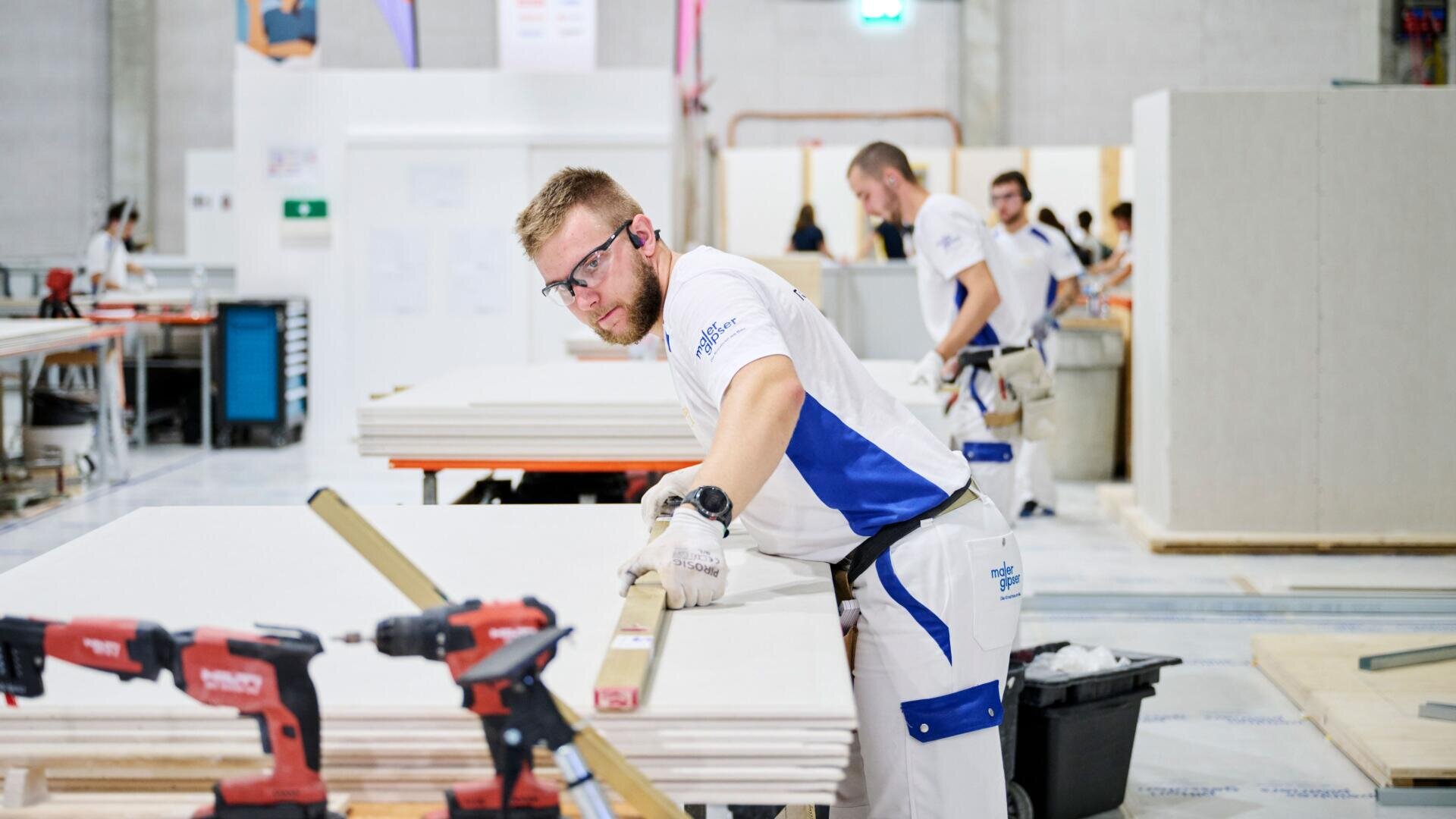 Ein Mann mit Schutzbrille und Handschuhen misst ein Blatt Material auf einer Werkbank in einer Werkstatt aus, während im Hintergrund Elektrowerkzeuge und andere Arbeiter zu sehen sind.